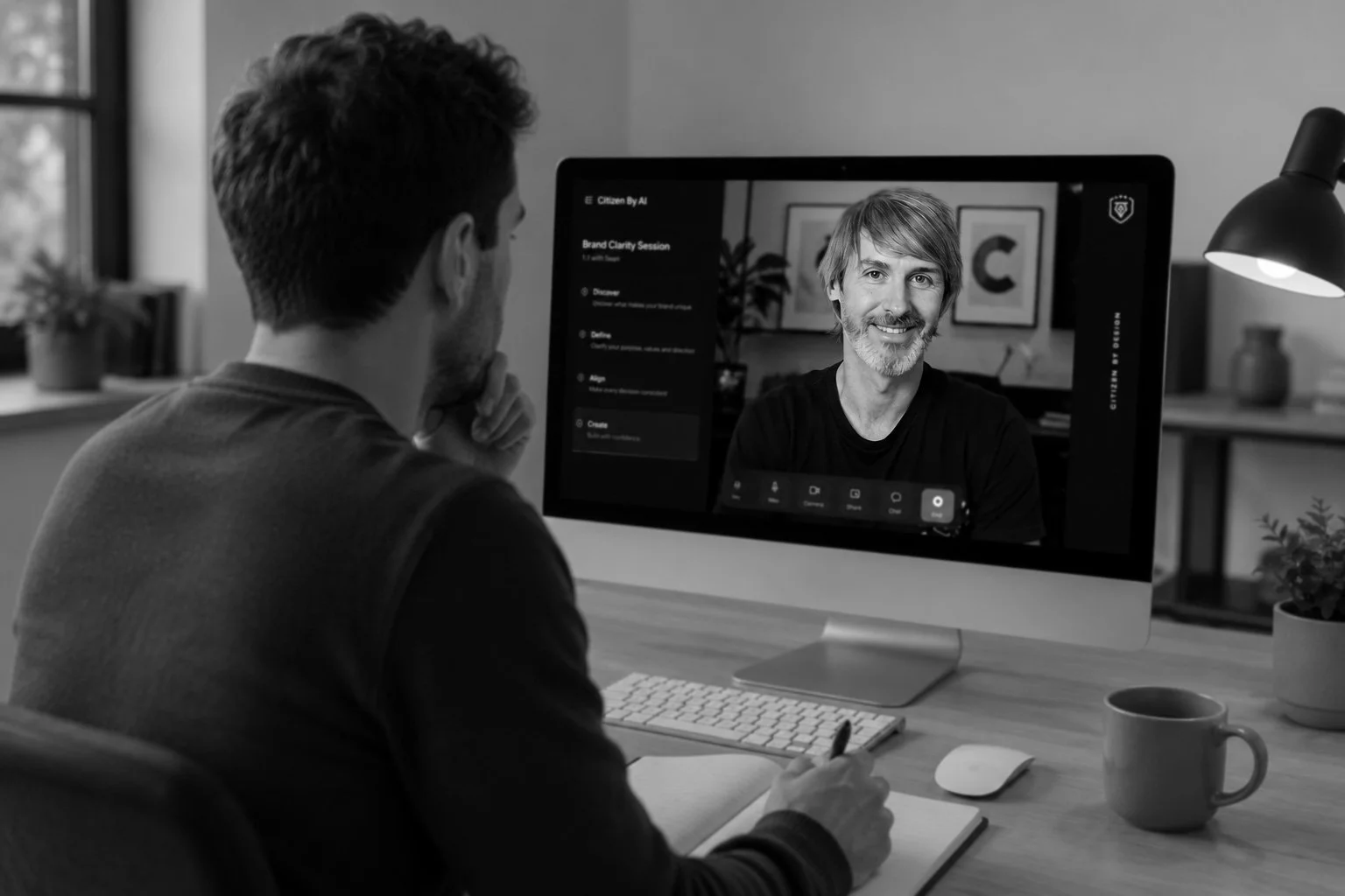A man sitting at a desk participating in a video conference on a computer, showing a smiling man on the screen.