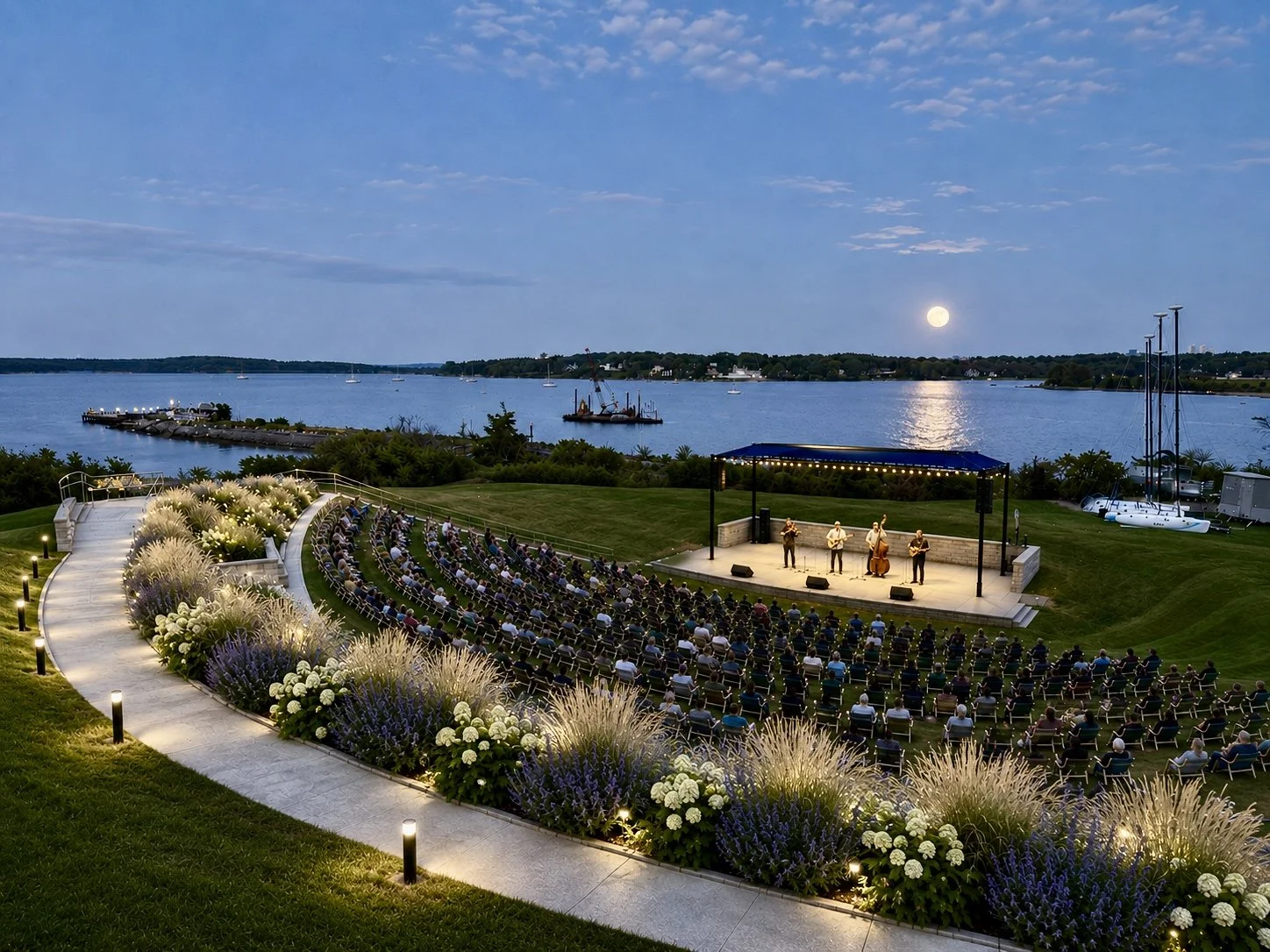 An outdoor concert near a body of water during dusk with people seated in rows facing a stage. Musicians perform under a canopy, illuminated by string lights, with boats and a full moon reflecting on the water.