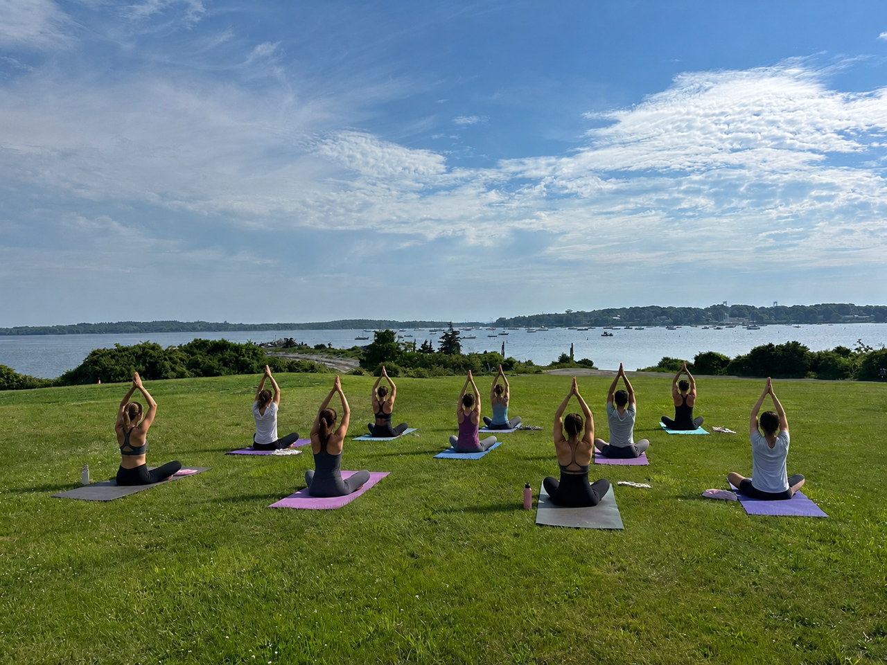 A group of ten people practicing yoga outdoors on a grassy field near a body of water, sitting on colorful yoga mats with their hands raised above their heads in a prayer position, under a partly cloudy sky.