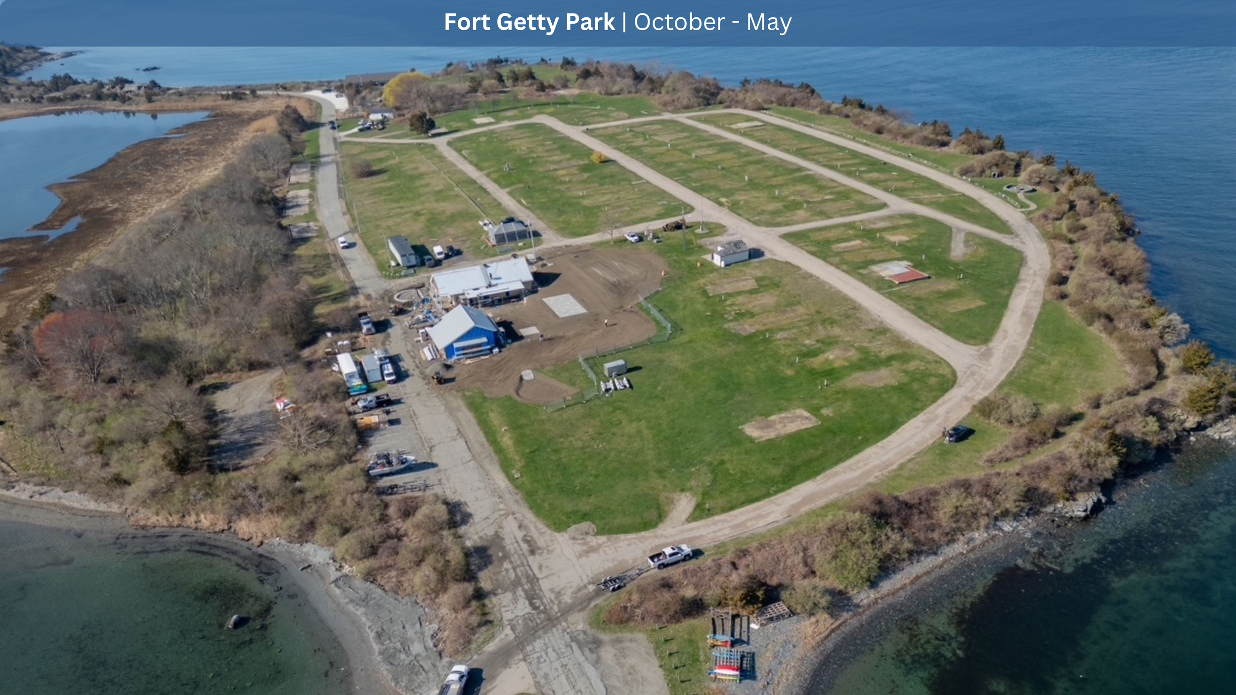 Aerial view of Fort Getty Park, with several open grassy areas, dirt roads, a few buildings and vehicles, surrounded by water on three sides, and some trees and a body of water to the west.