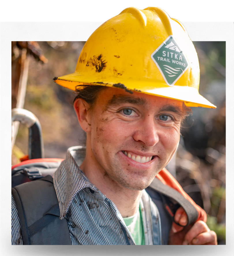 A smiling man wearing a yellow hard hat with a "Sitka Trail Works" logo, and a backpack, outdoors on a trail.