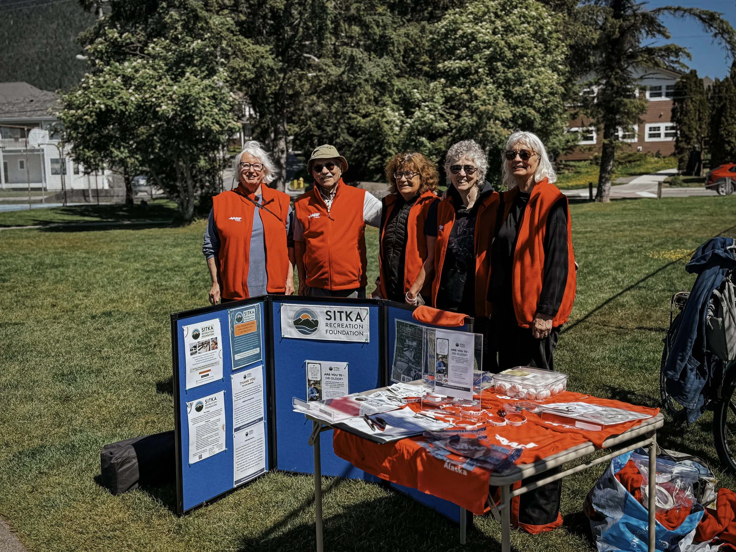 A group of five elderly people wearing orange vests stands behind an outdoor display table at a park or fair, with informational posters and branded merchandise, in front of trees and houses on a sunny day.