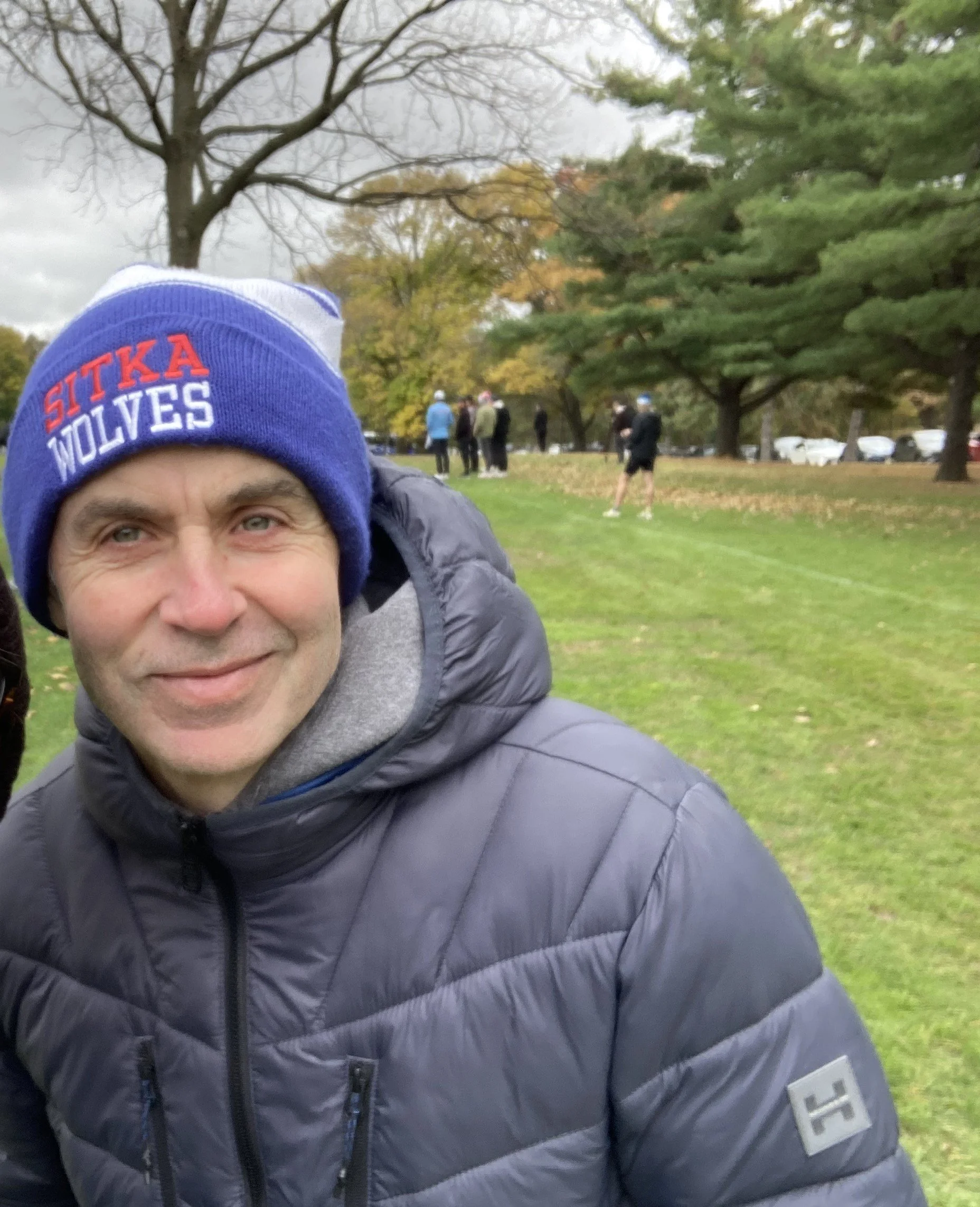 Close-up of a man's face wearing a blue beanie that says 'SITKA WOLVES', in a park with trees and people in the background.