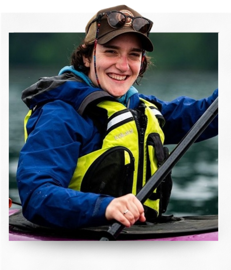 Smiling woman in a yellow life jacket and blue waterproof jacket kayaking on a body of water during daytime.