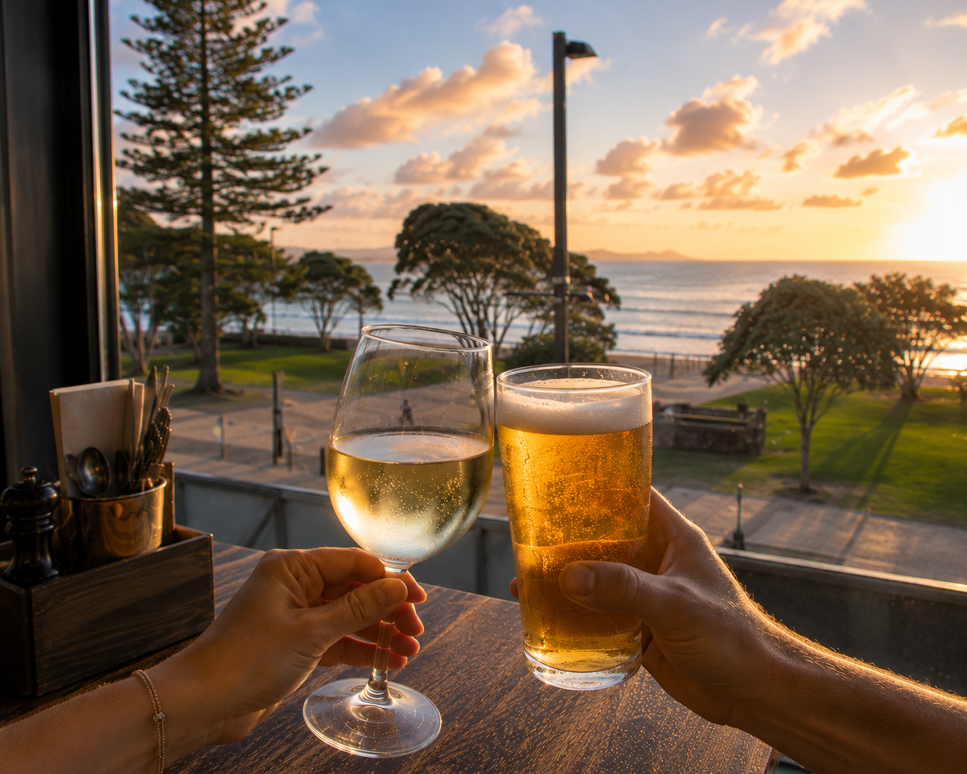 Two hands holding a glass of white wine and a glass of beer with a sunset view of trees, the ocean, and a partly cloudy sky in the background.