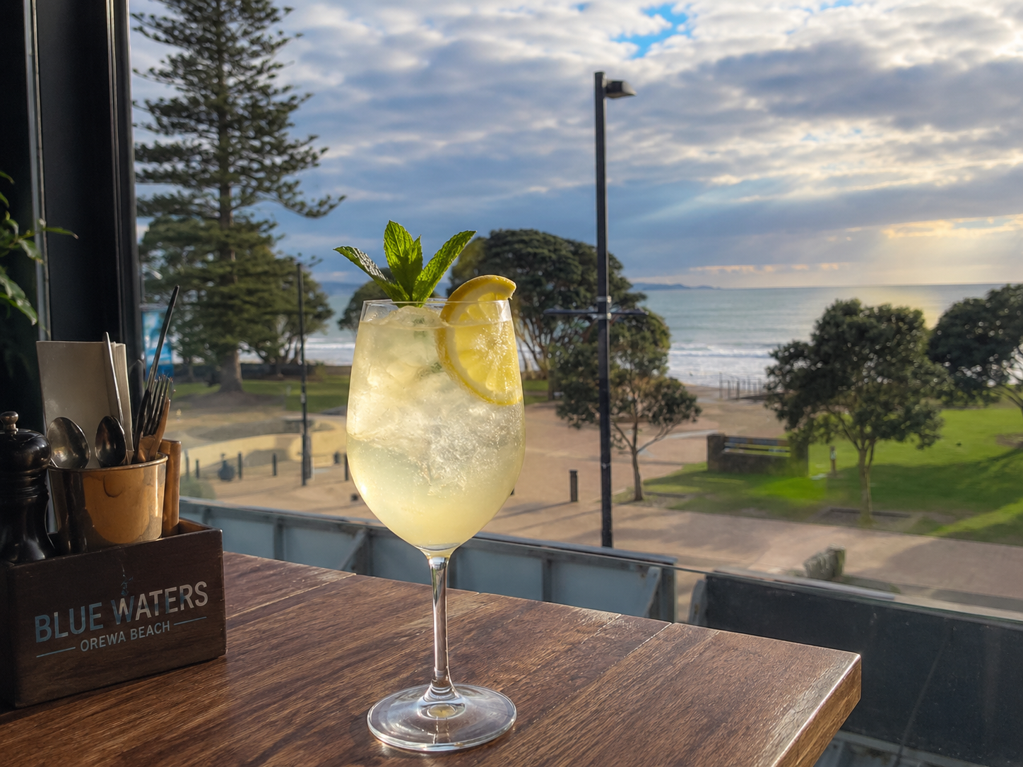 A glass of lemonade garnished with lemon slices and mint leaves on a wooden table overlooking a beach with trees, sand, and the ocean at sunset.