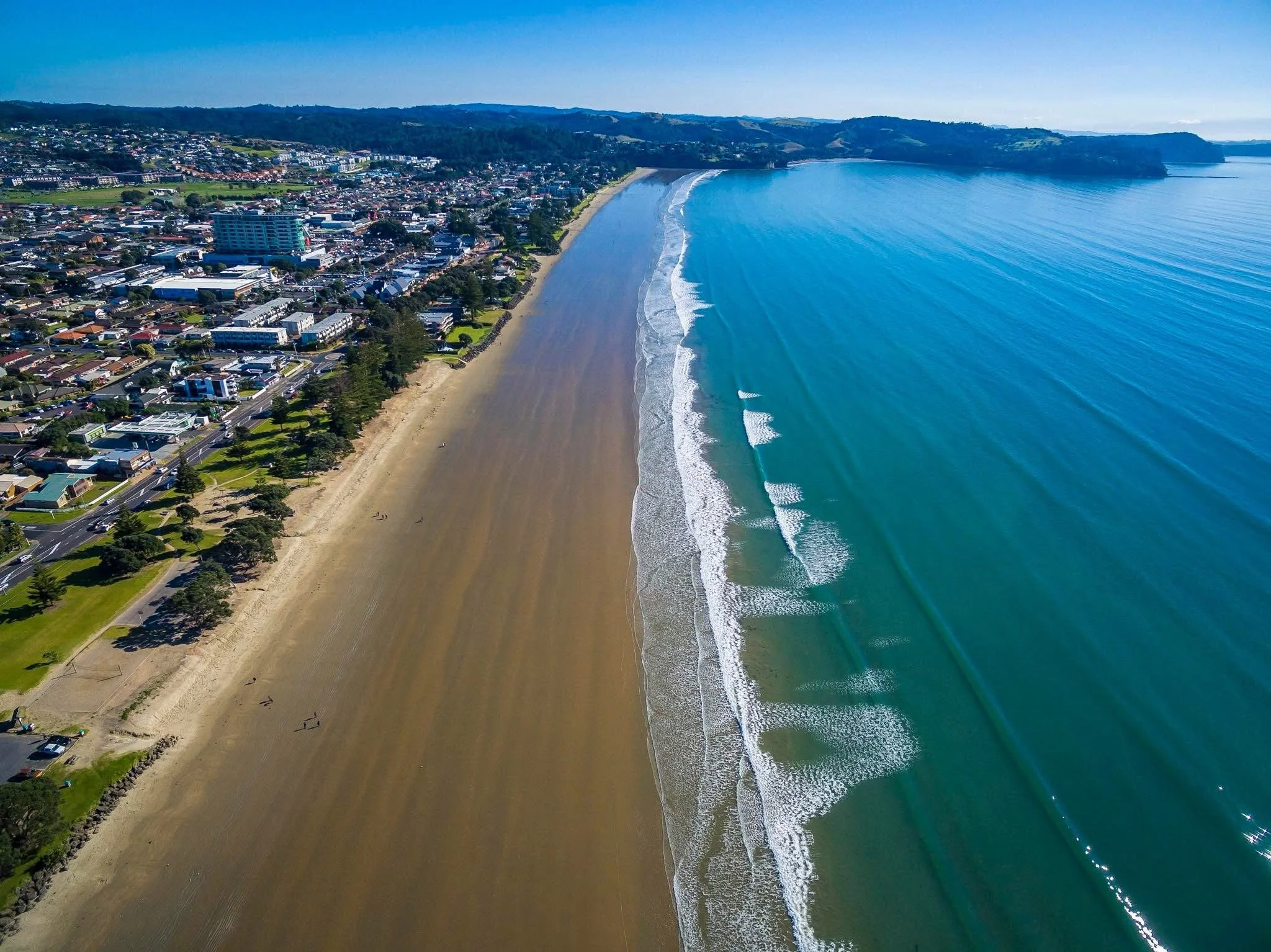 Aerial view of a coastal city with a beach, ocean waves, and urban area with buildings and roads.