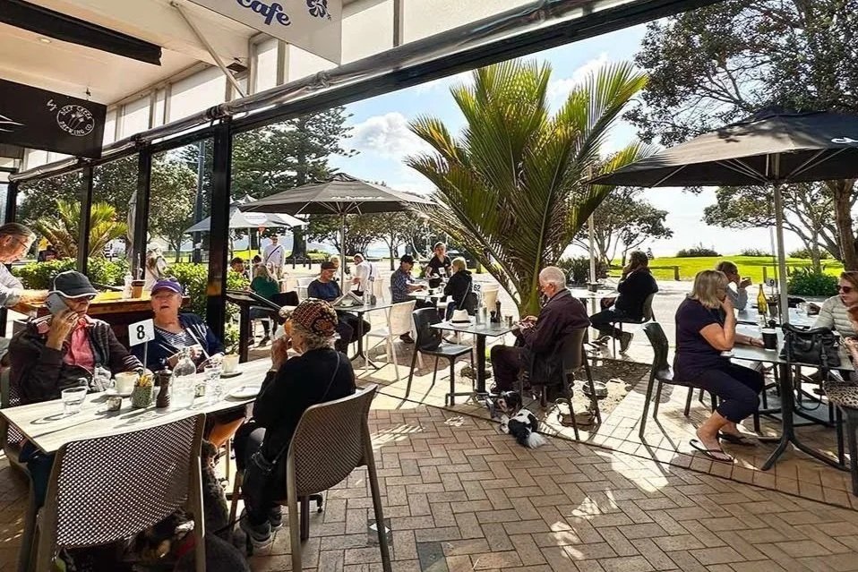 People dining outdoors at a cafe with umbrellas, palm trees, and a grassy area in the background.