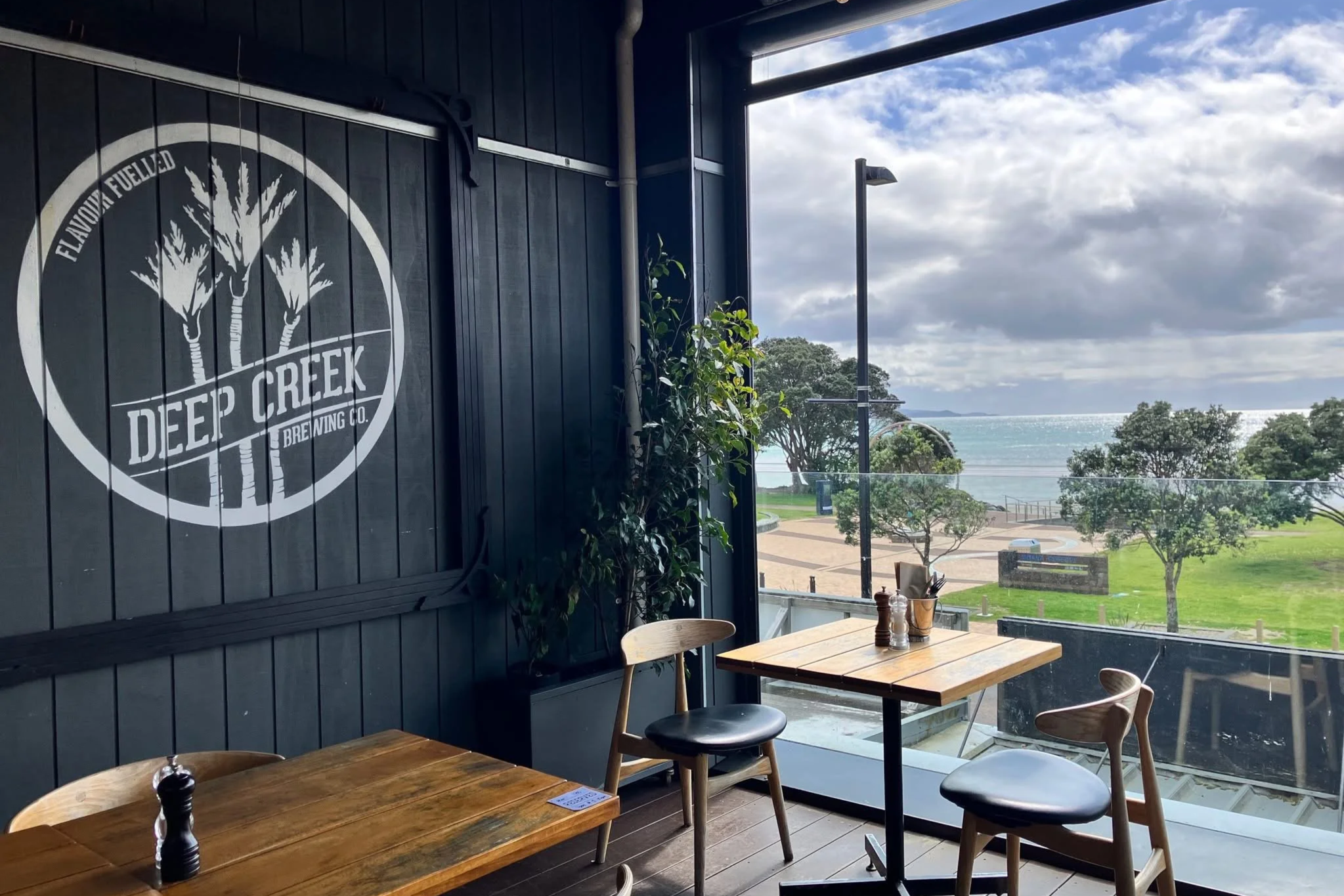 Indoor seating area of a cafe with wooden tables and chairs, a dark wall with a White Deep Creek Brewing Co. logo, large windows showing a park, trees, and the ocean under a partly cloudy sky.