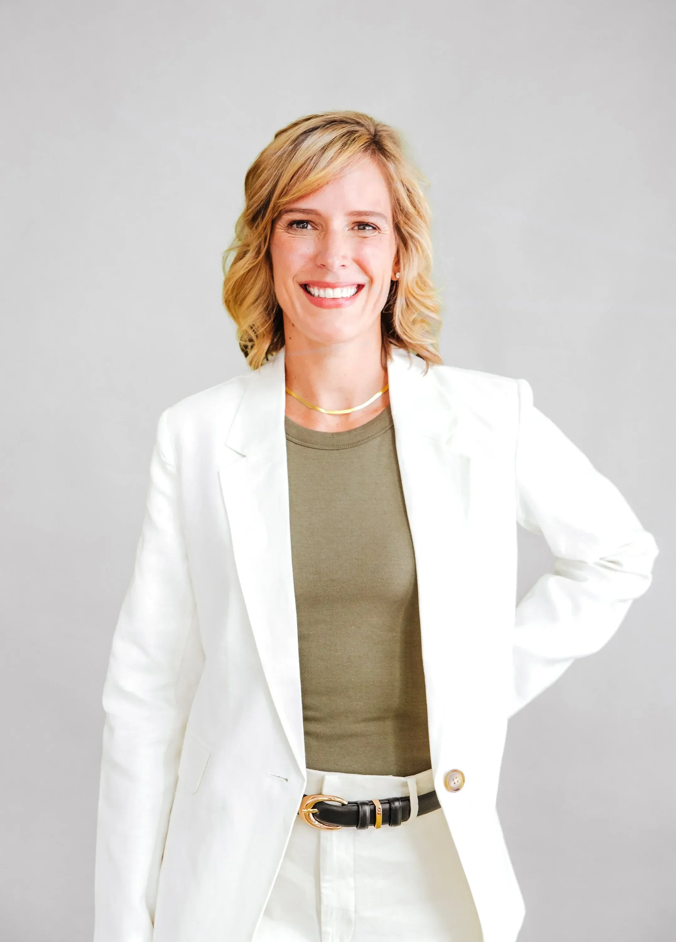 Dr. Ashley Chambers with blonde wavy hair smiling, wearing a white blazer, green top, and a black belt with gold accents, standing against a plain light gray background.