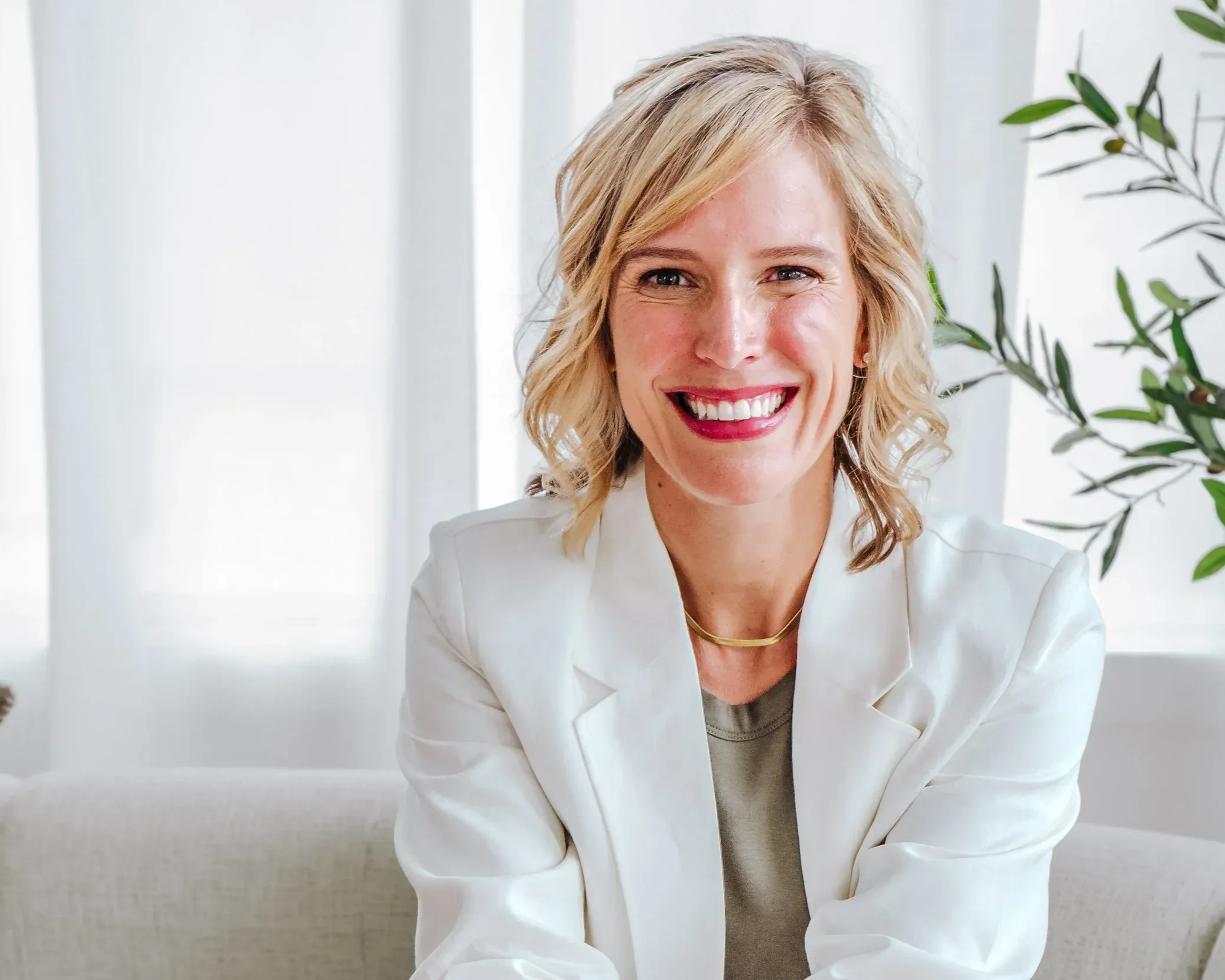 Dr. Ashley Chambers wearing a white blazer and light-colored top, sitting in a bright, airy room with white vertical blinds and green leafy plants in the background.