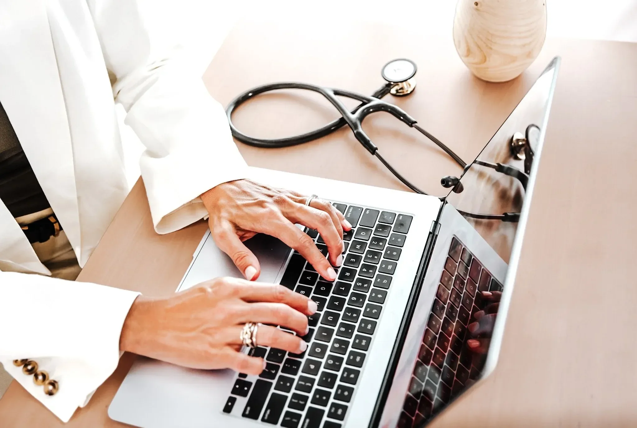 Close-up of a person typing on a laptop keyboard with a stethoscope and a vase on the table.