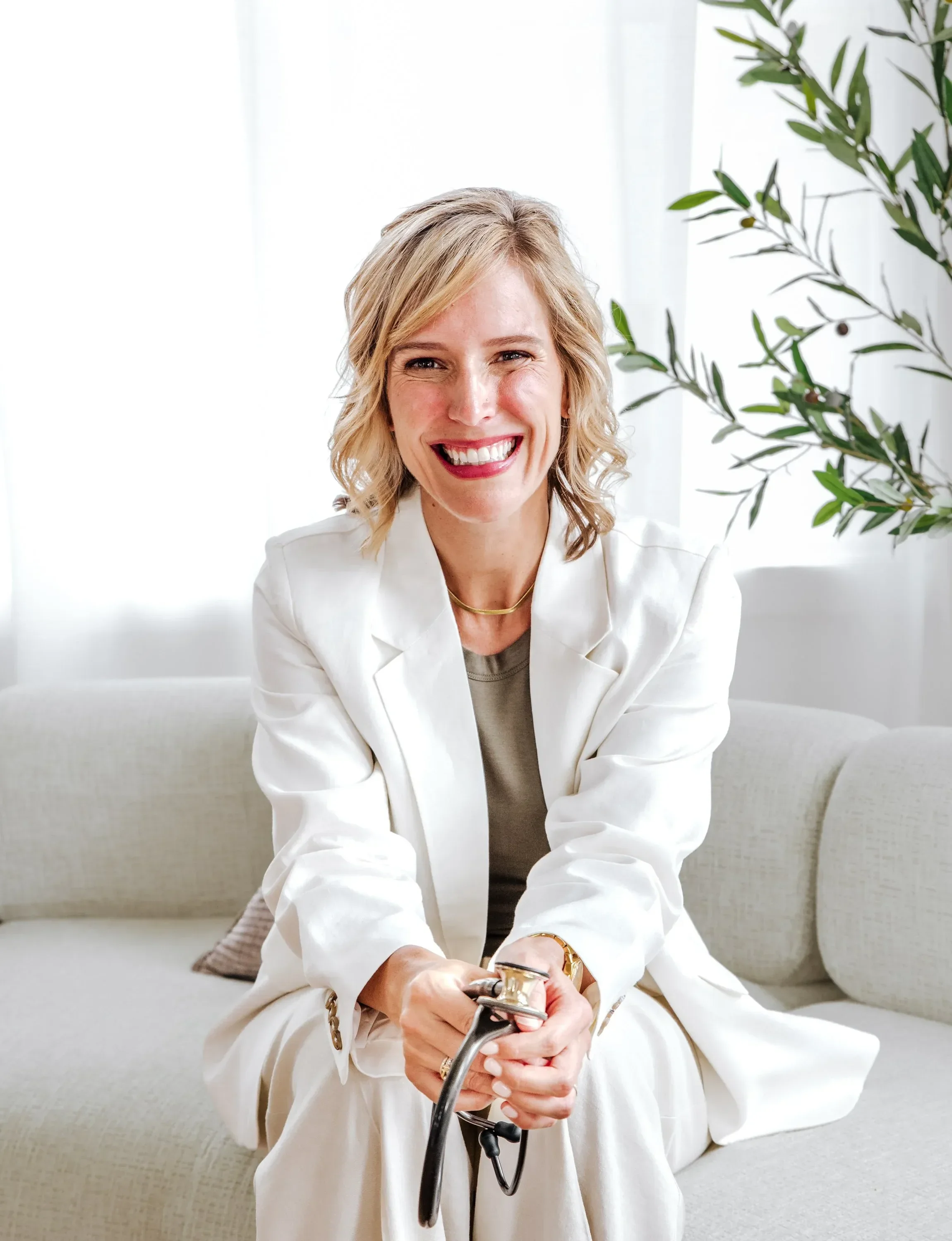 Dr. Ashley Chambers smiling brightly while sitting on a light-colored sofa, holding a stethoscope, in a room with greenery and white curtains.