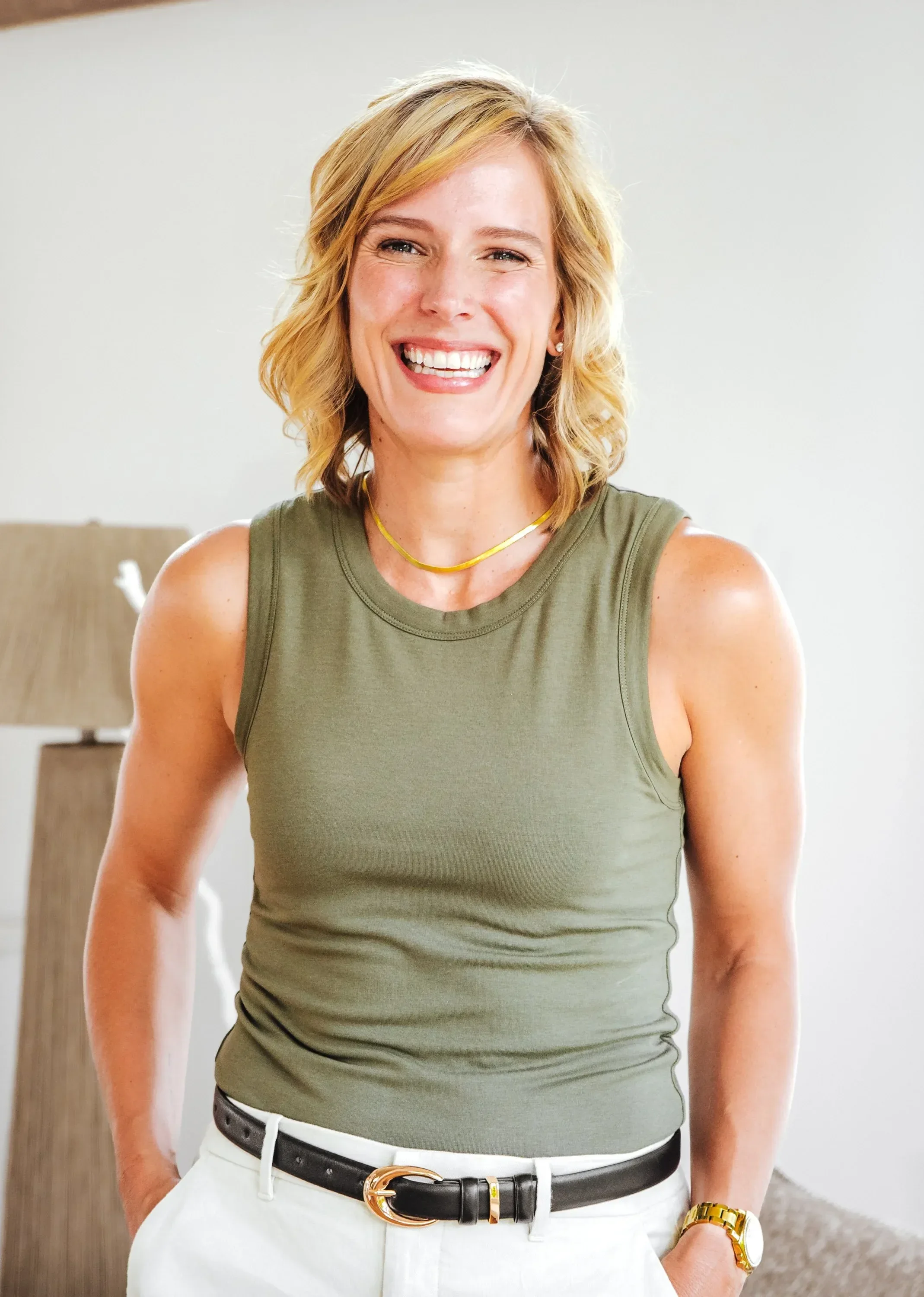 Dr. Ashley Chambers with shoulder-length blonde hair smiling, wearing a green sleeveless top, white pants, a black belt with a gold buckle, a gold watch, and a yellow necklace.