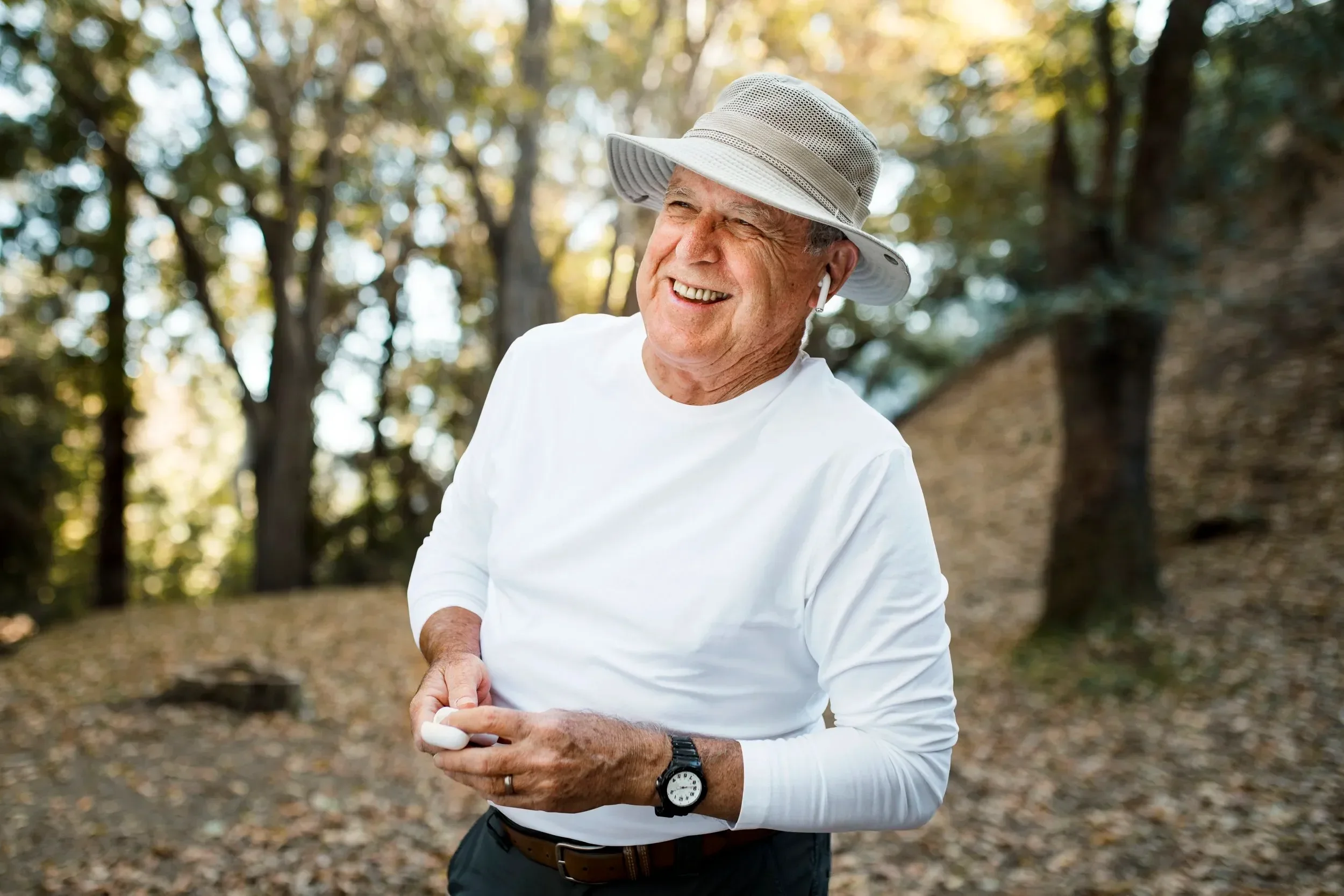 Smiling elderly man in sunglasses and hat, wearing a white shirt, stands in a sunlit forest, conveying joy and serenity while holding a small device.
