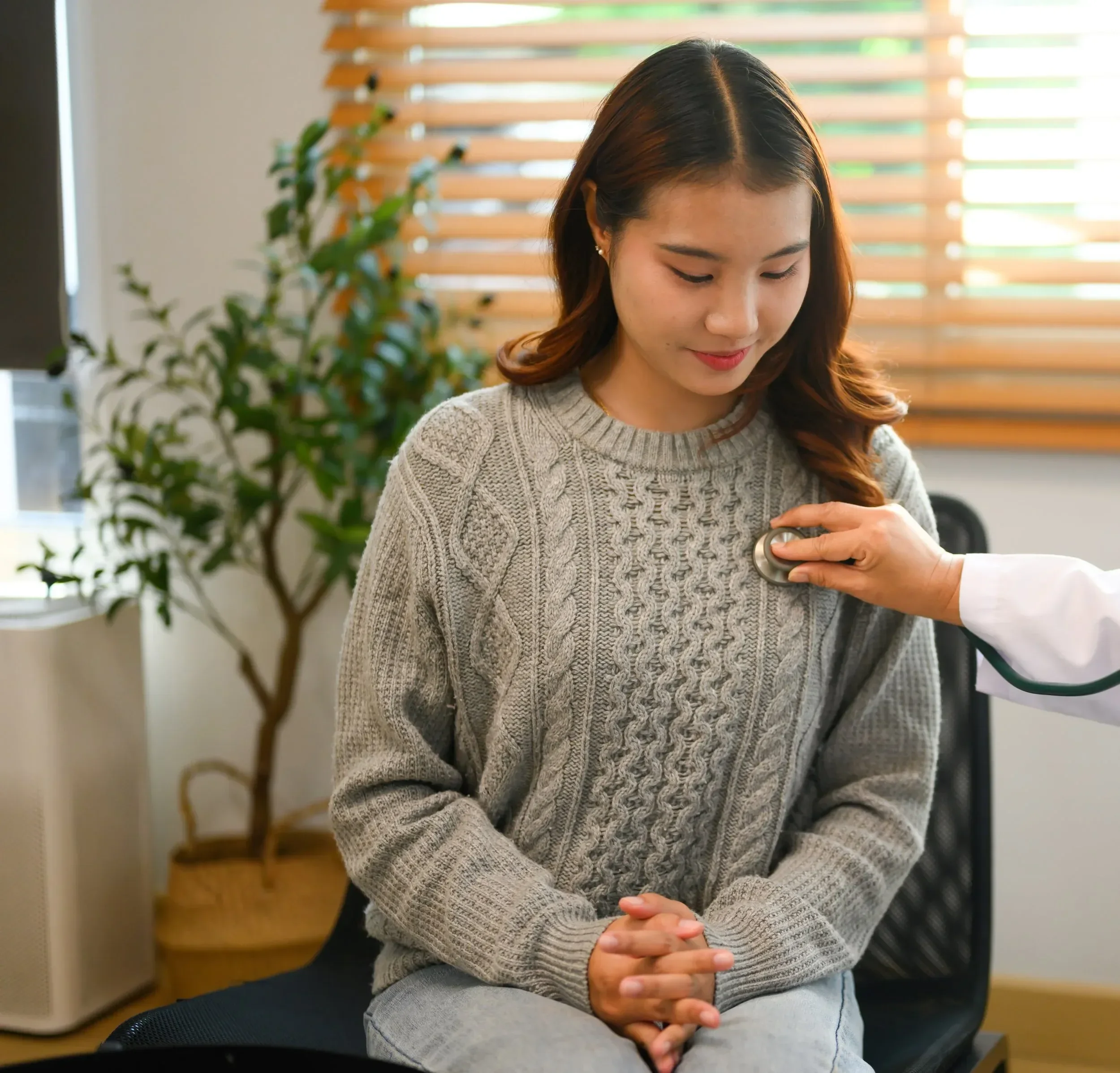 A woman in a gray sweater sits calmly while a doctor's hand places a stethoscope on her chest. The room is bright with a plant in the background.