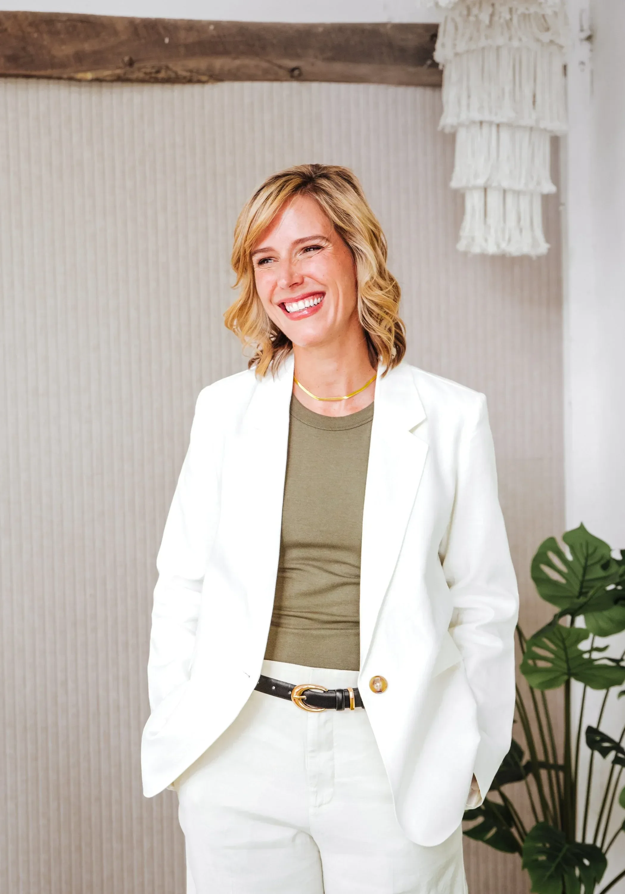 Dr. Ashley Chambers with blonde, wavy hair wearing a white blazer, light green shirt, and white pants, standing indoors with a wooden ceiling, decorative hanging lamp, and a plant in the background.