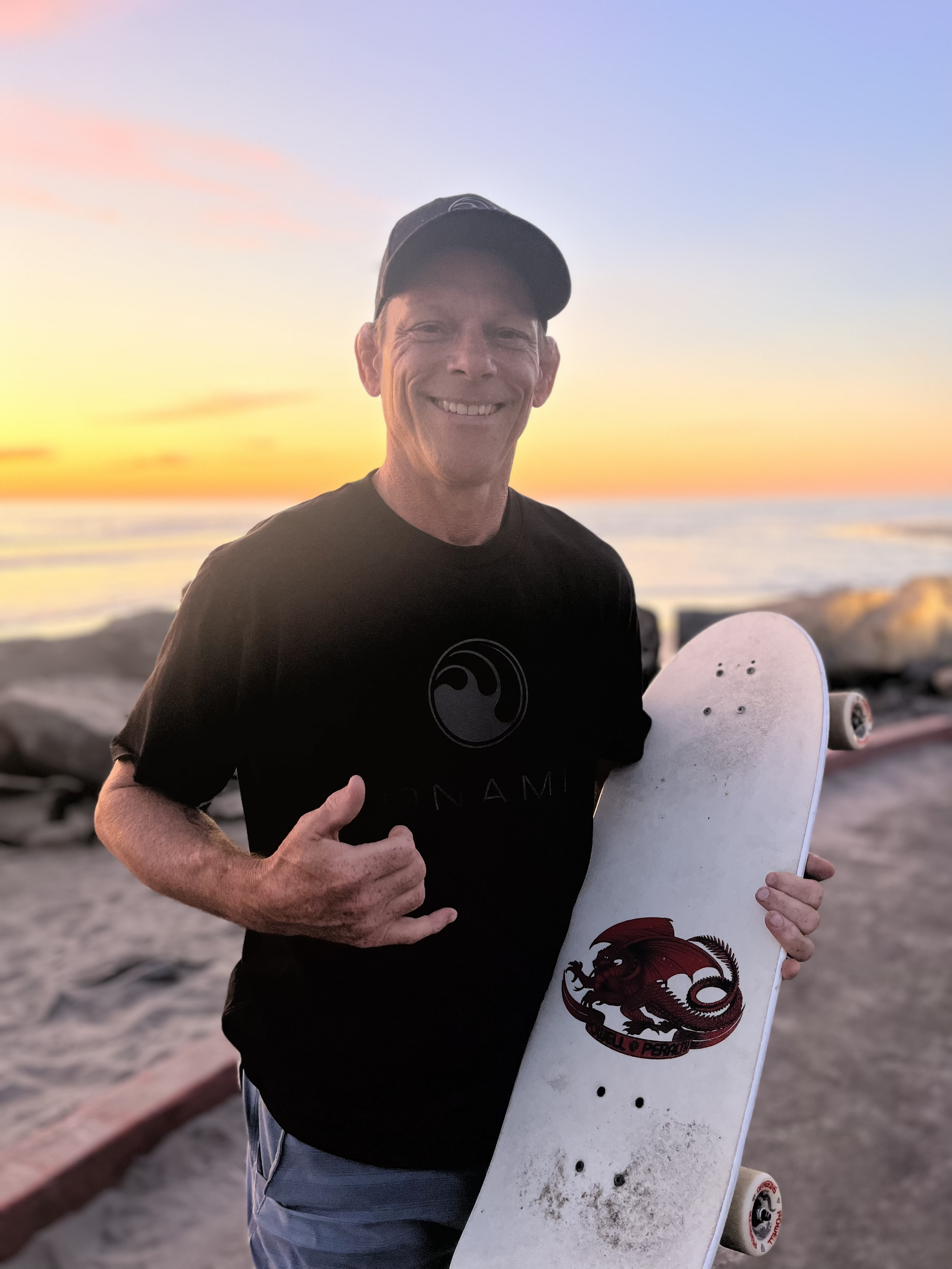 A man smiling and giving a thumbs-up while holding a skateboard with a dragon graphic at the beach during sunset.