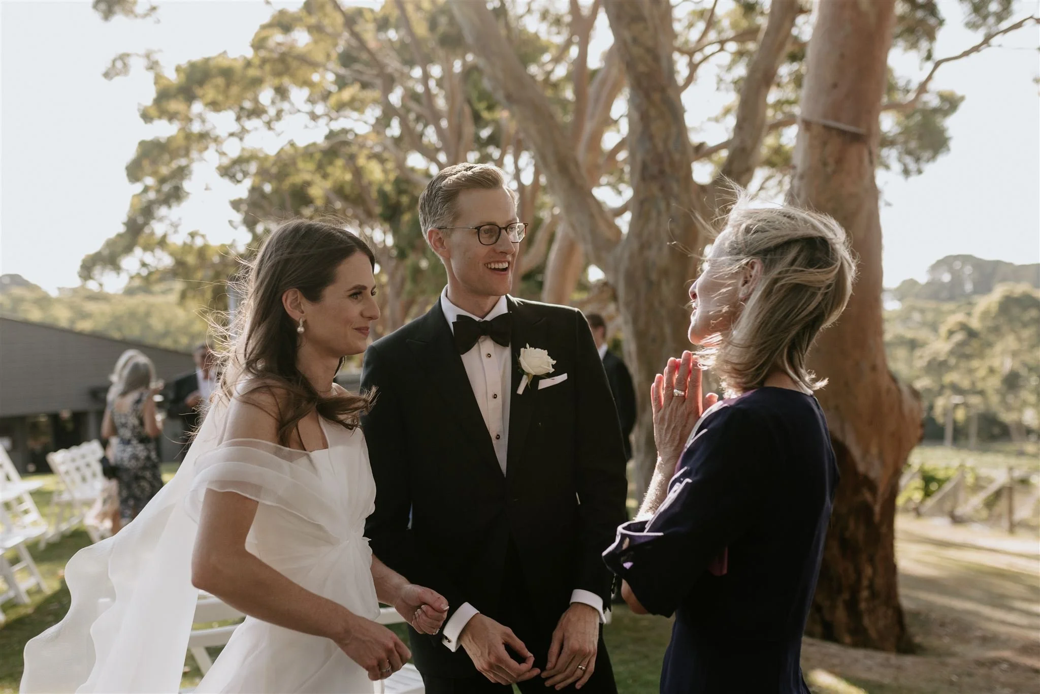 A bride and groom holding hands and smiling at each other during a wedding ceremony outdoors, with a woman in a dark dress facing them. The background features trees and chairs.