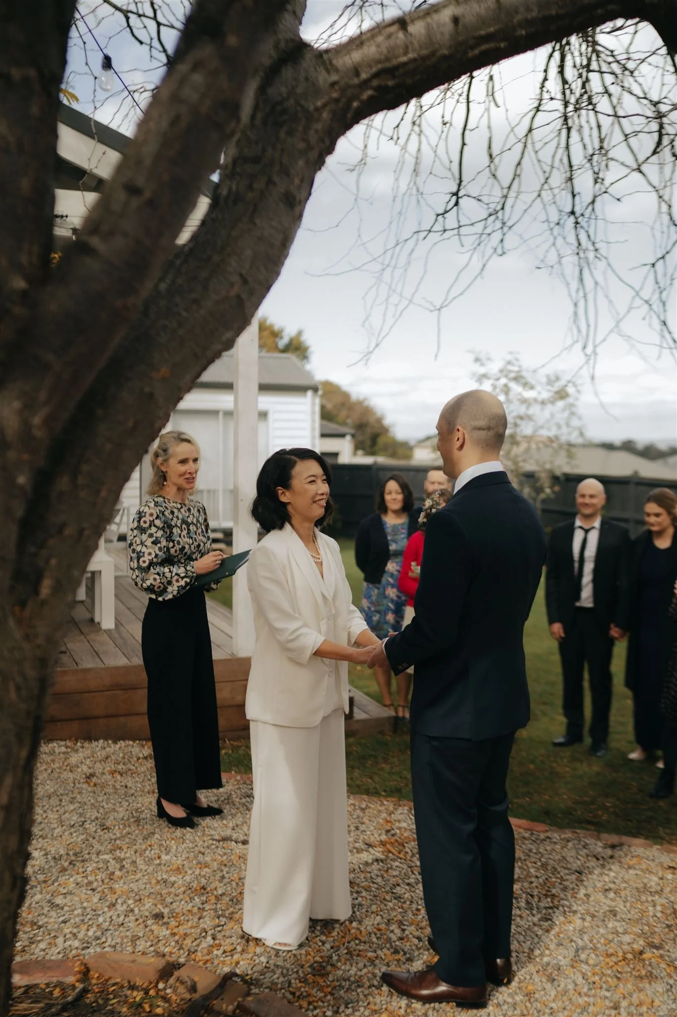 A wedding ceremony outdoors, with a woman in a white suit and a man in a dark suit holding hands and exchanging vows. Several onlookers are standing in the background, dressed in formal and semi-formal attire. The scene is framed by leafless tree branches.
