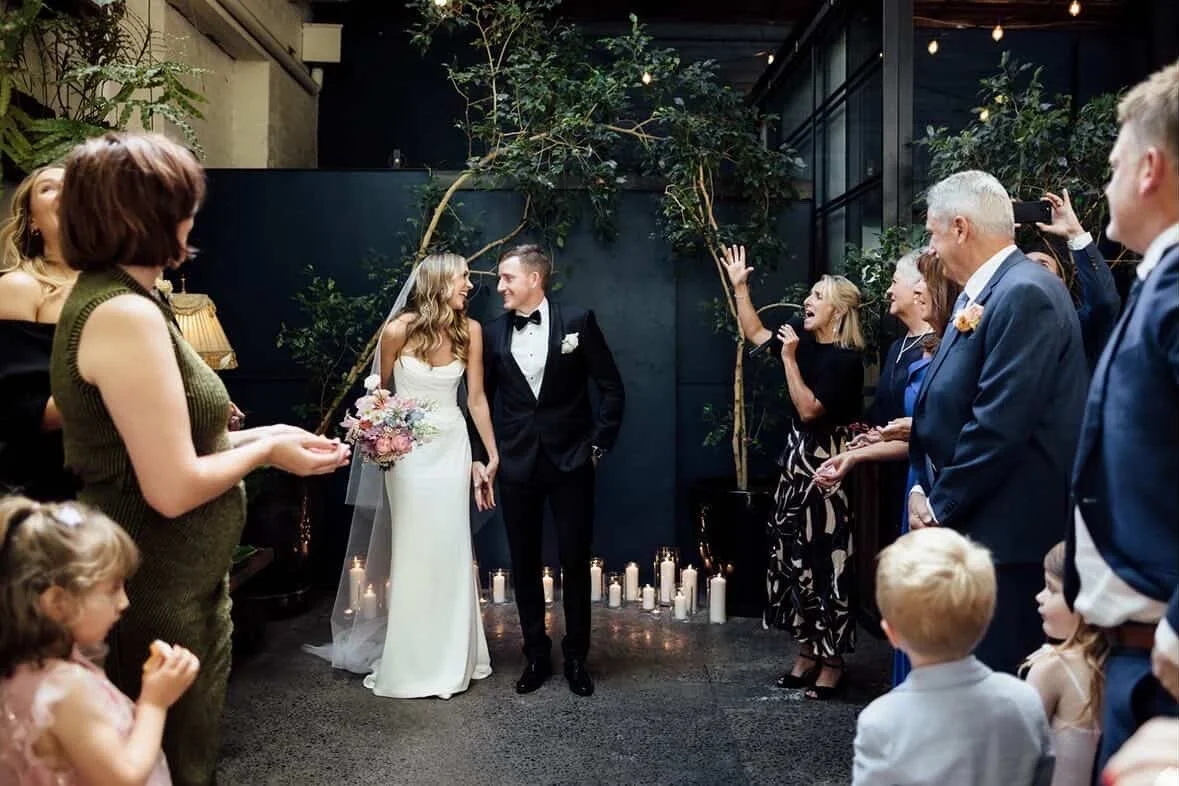 A bride and groom stand close together, holding hands and smiling at each other during their wedding ceremony. They are surrounded by family and friends who are celebrating and taking photos indoors with candles and plants in the background.
