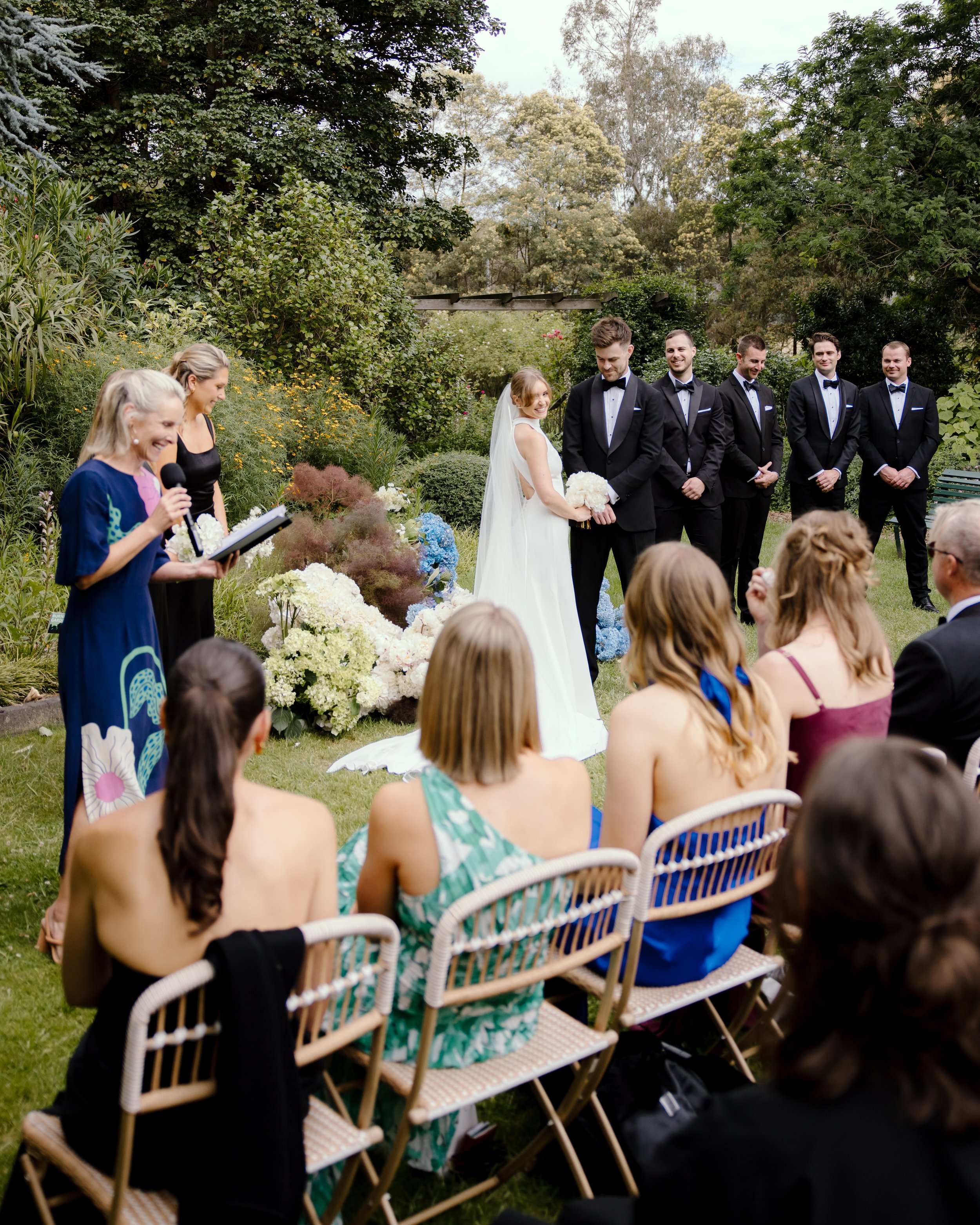 A wedding ceremony outdoors with the bride and groom in the center, surrounded by guests seated on chairs. The bride is holding a bouquet and smiling at the groom, who is dressed in a tuxedo. Bridesmaids and groomsmen are lined up behind them, all dressed formally. The setting is lush with greenery and flowers.