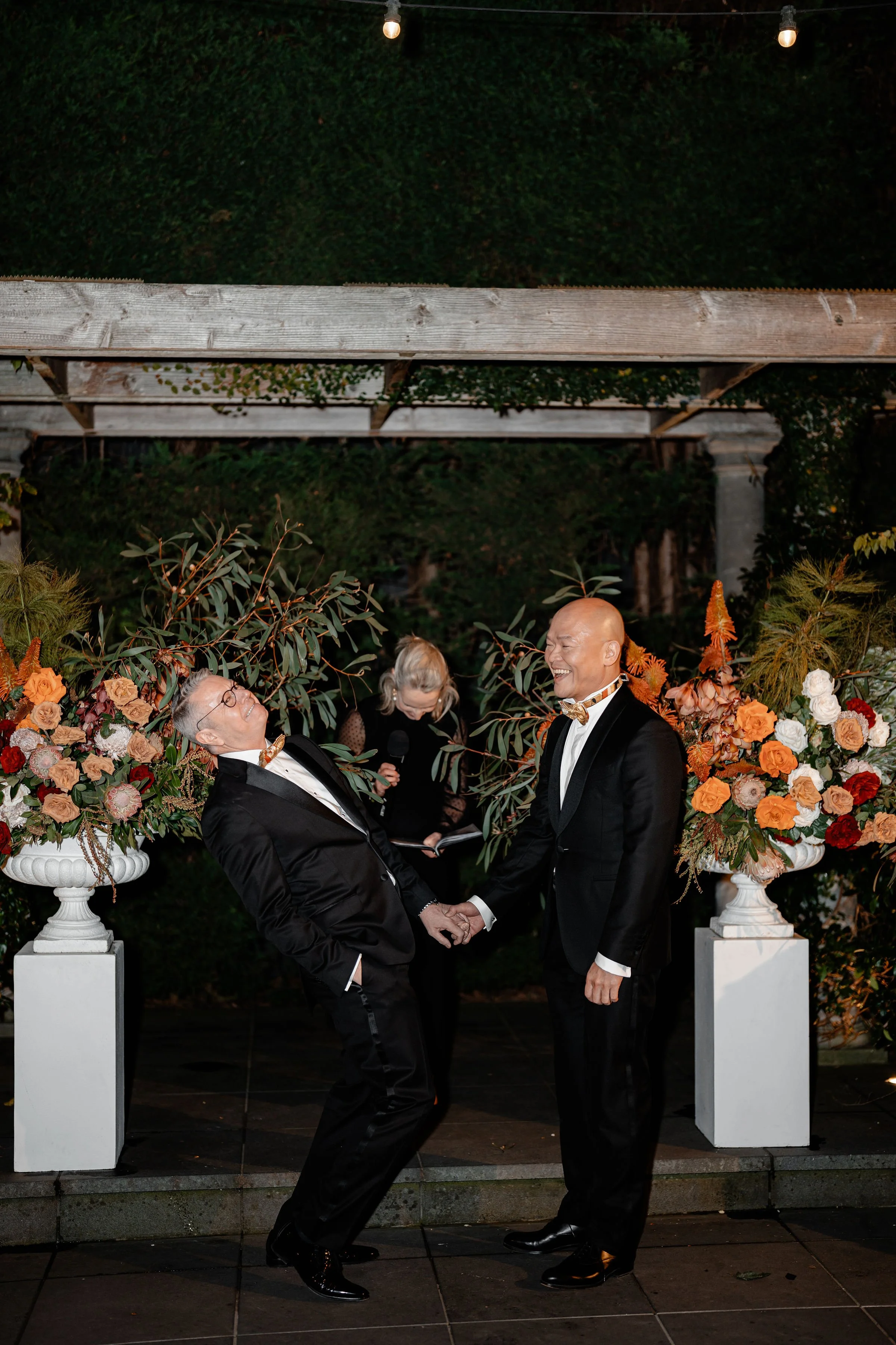 Two men in tuxedos holding hands and smiling during a wedding ceremony outdoors at night. They are surrounded by large floral arrangements and standing on a tiled platform with greenery and string lights overhead.