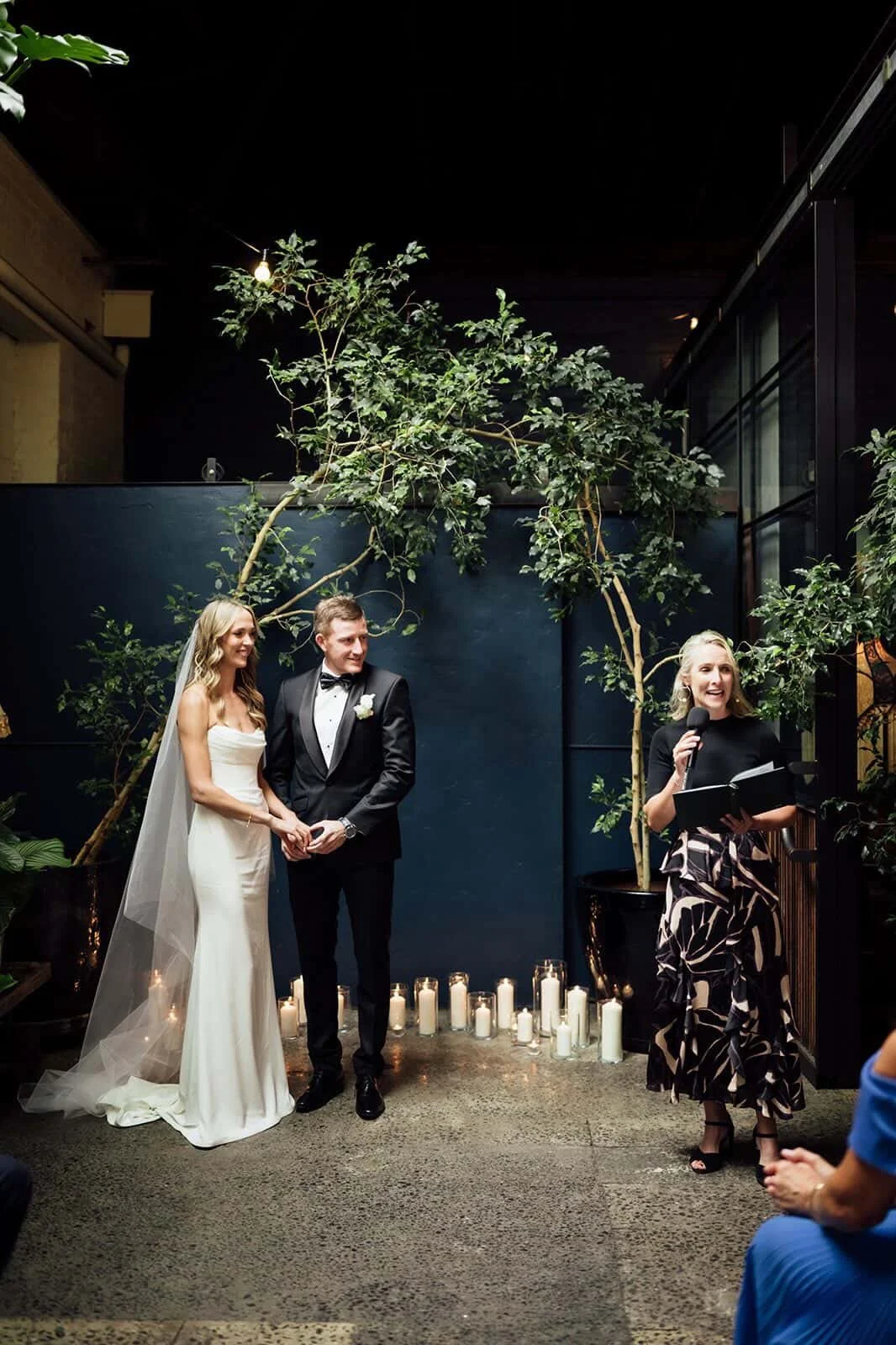 Bride and groom standing together during a wedding ceremony, listening to a woman speaking into a microphone, with candles and greenery in the background.