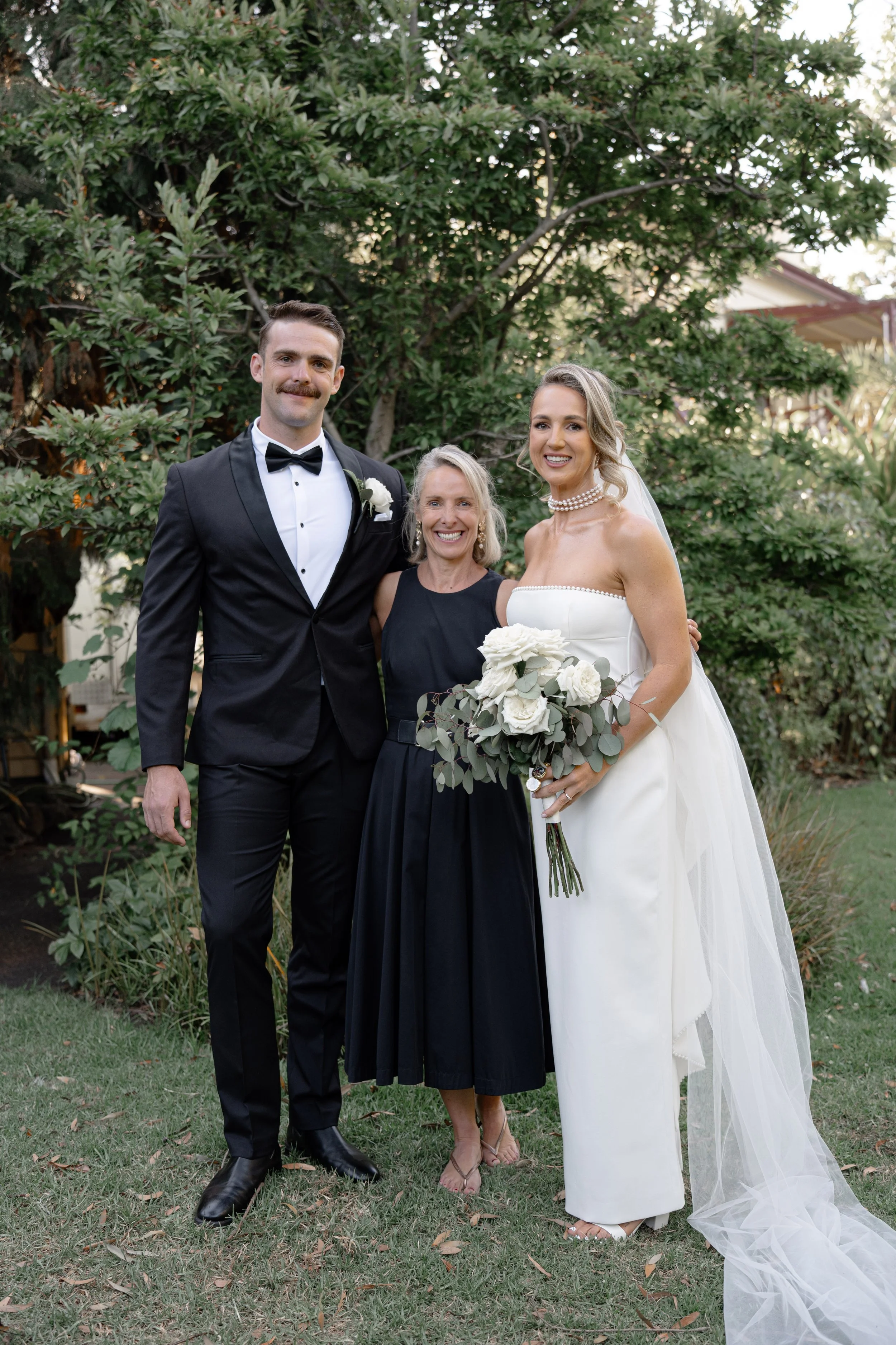 A bride in a white wedding dress holding a bouquet of white roses, a groom in a tuxedo, and a woman in a black dress standing outdoors in front of green trees.