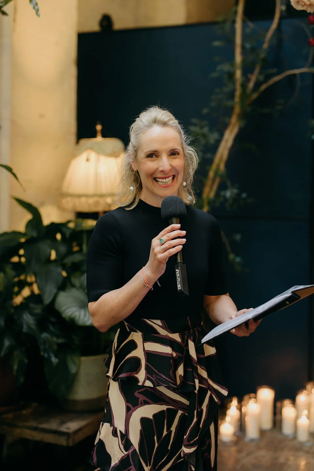 A woman with blonde hair smiling while holding a microphone and a tablet in a cozy, dimly lit indoor space decorated with plants, candles, and a lamp.