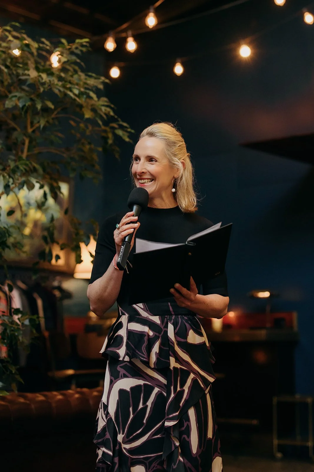 A woman with blonde hair holding a microphone and a black folder, smiling while speaking at an indoor event with string lights overhead.