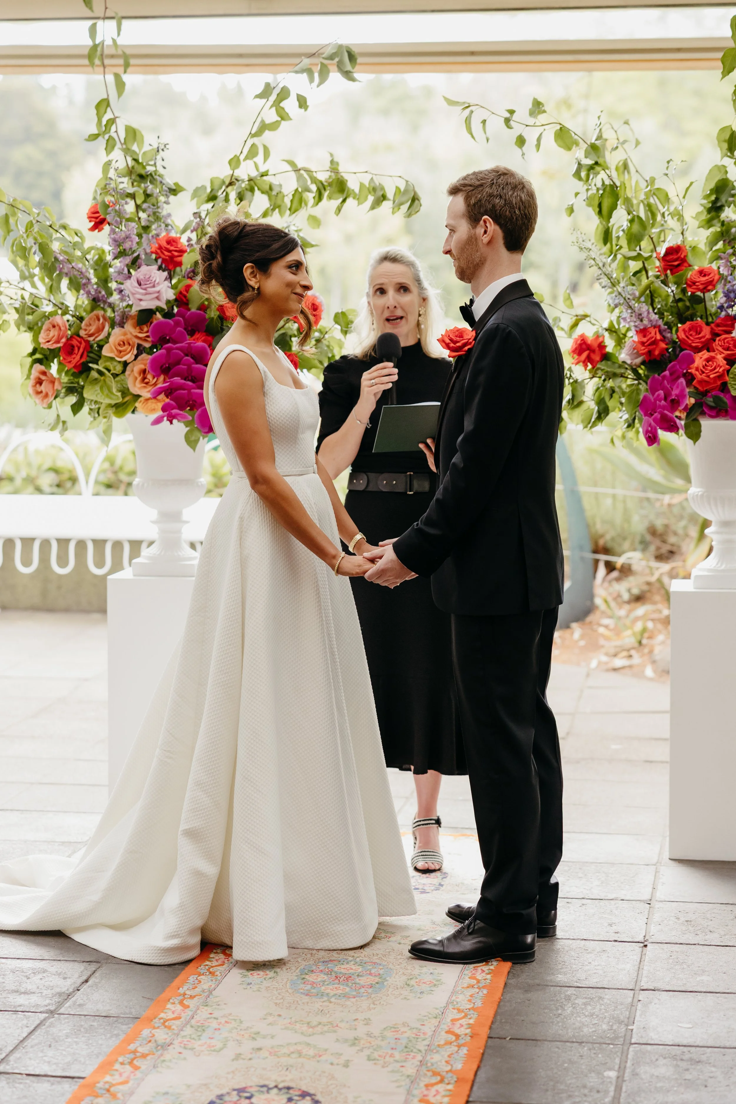 A bride and groom standing face to face during their wedding ceremony, holding hands, with an officiant speaking in the background. The bride is wearing a white wedding dress and the groom is in a black tuxedo. The setting is decorated with large floral arrangements and a patterned rug.