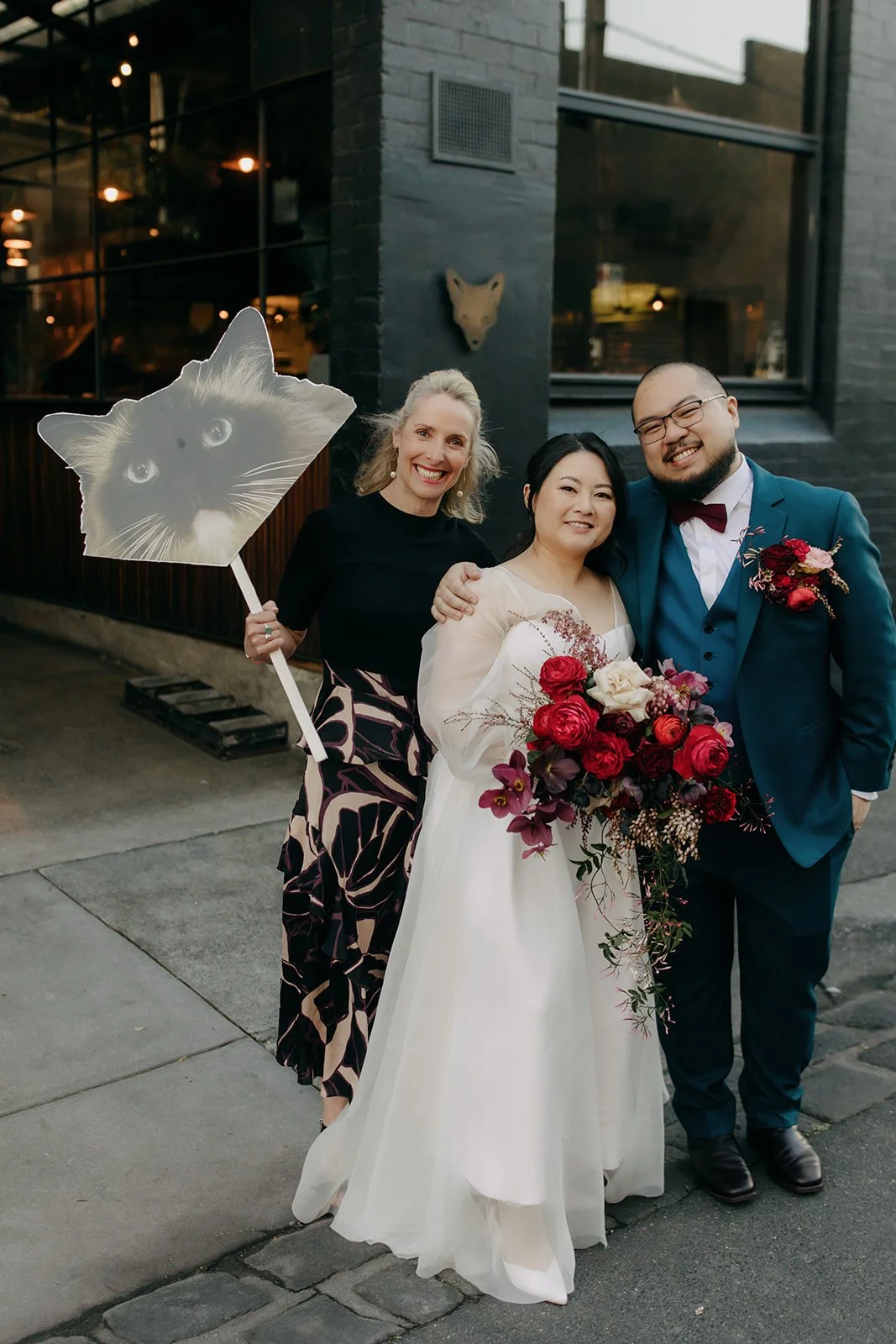 A bride and groom smiling with two women, one holding a cat-themed sign, outside a modern building with black brick walls and large windows at night.