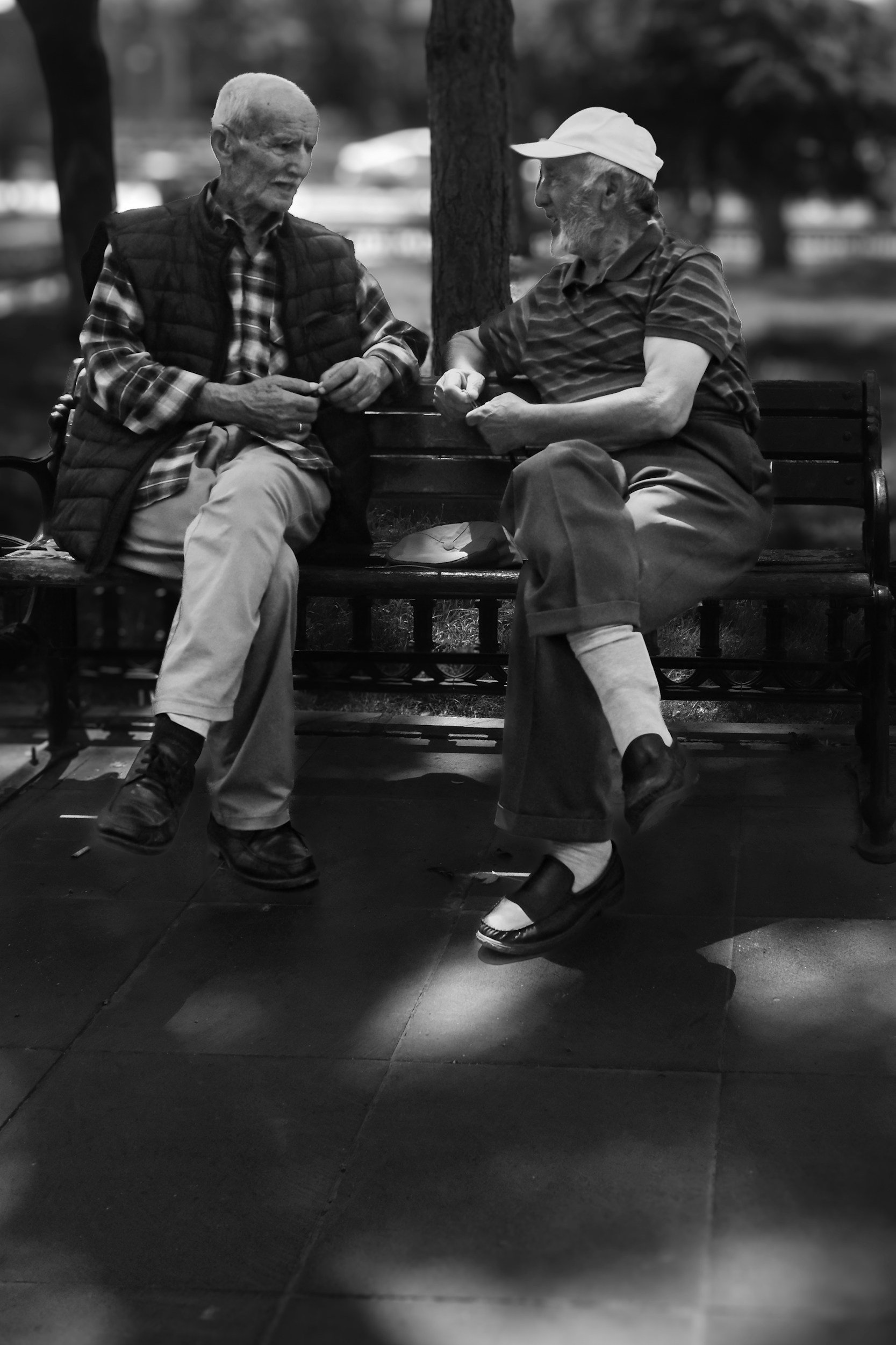 Two elderly men are sitting on a park bench, engaged in conversation. One is wearing a plaid shirt and a quilted vest, while the other is dressed in a striped shirt, shorts, and a baseball cap. They appear to be enjoying a friendly chat outdoors.