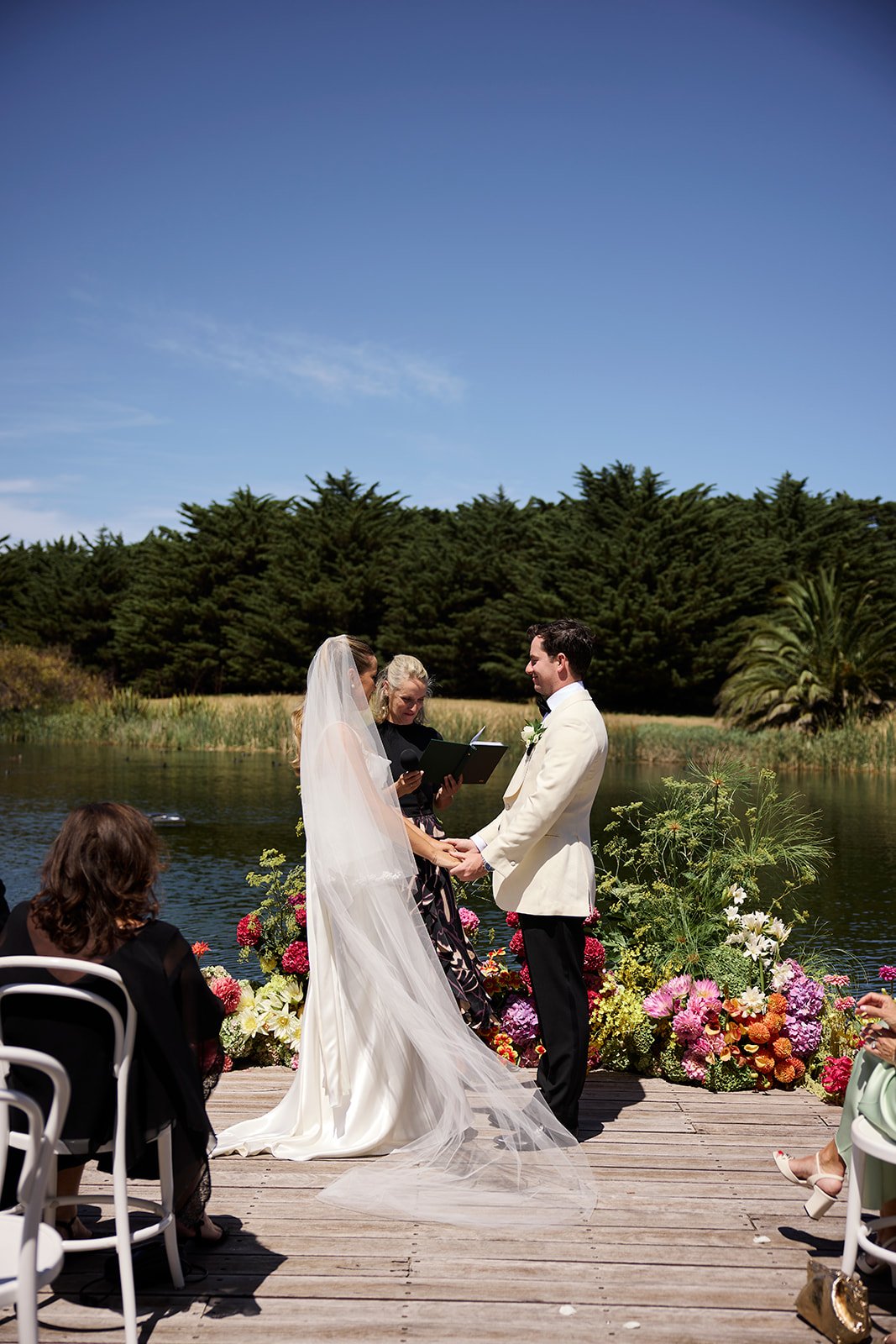A wedding ceremony taking place outdoors on a wooden dock by a lake, with a bride and groom holding hands, an officiant reading, and guests seated nearby, surrounded by colorful flowers and lush green trees under a blue sky.