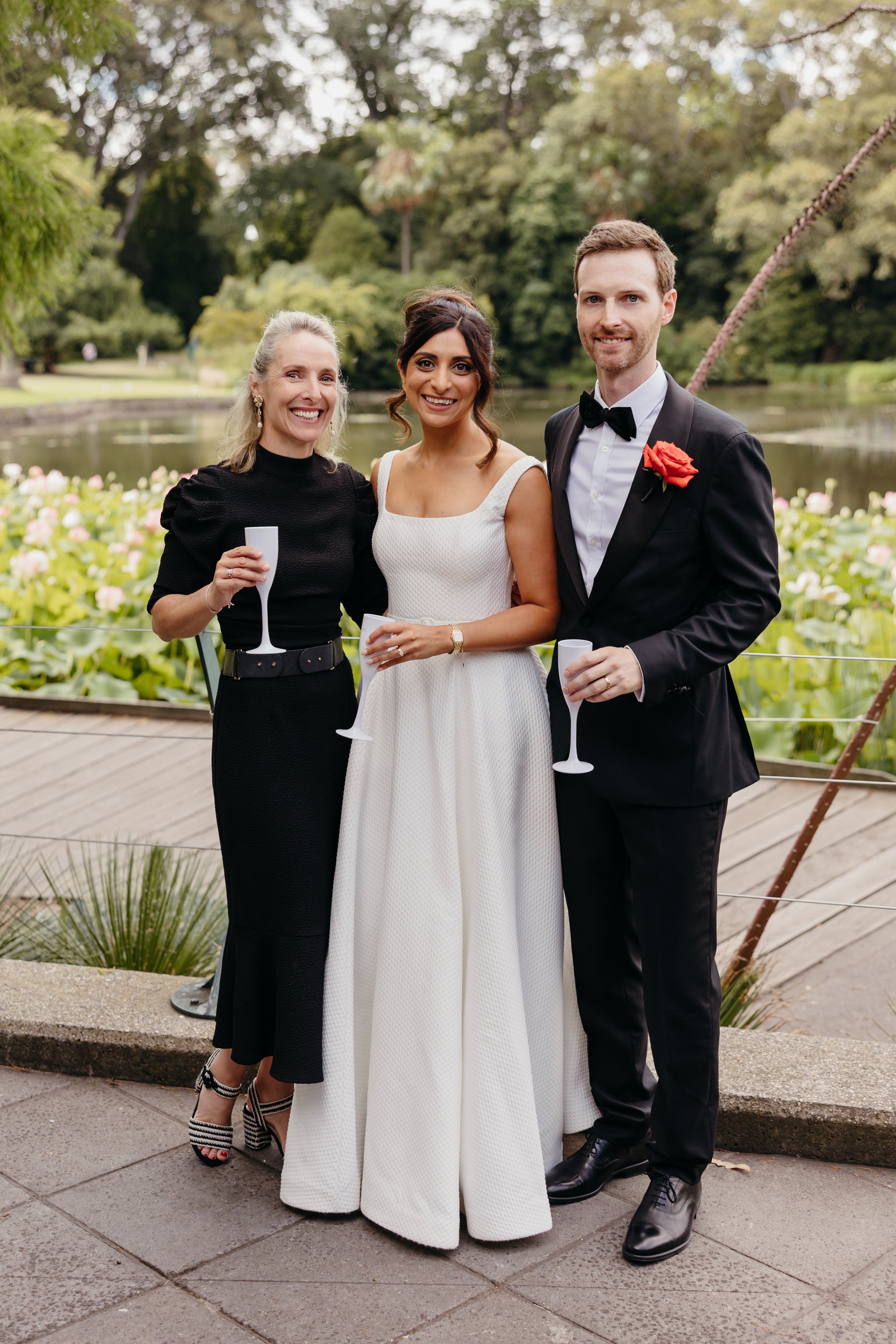 A group of three people, two women and one man, smiling and holding champagne glasses, standing outdoors near a pond and surrounded by greenery, during a celebration or special event.