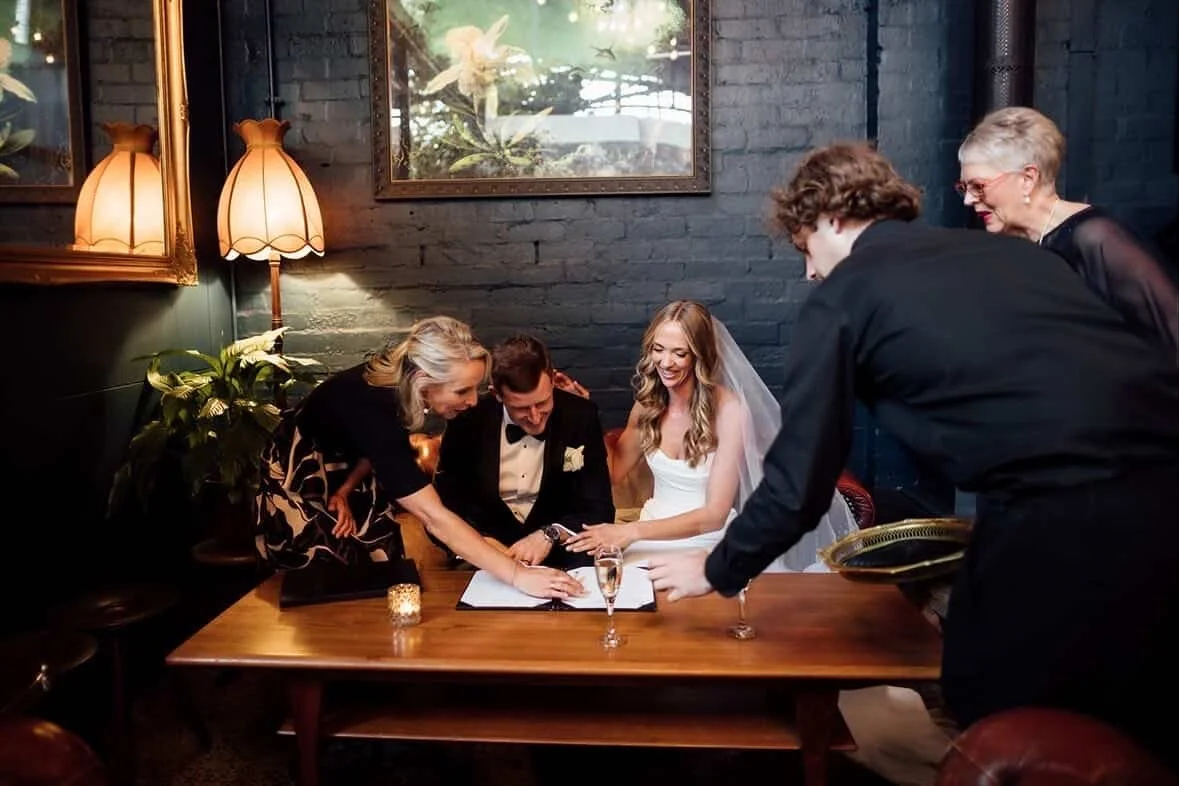 A wedding celebration with the bride, groom, and guests signing a guest book at a table in a cozy indoor setting.