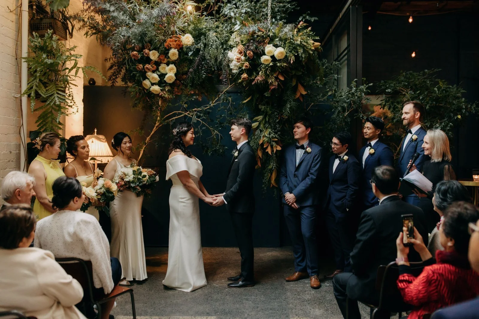 A wedding ceremony where a bride and groom stand facing each other, holding hands, in front of a floral arch. They are surrounded by bridesmaids and groomsmen, with guests seated and watching. The setting is decorated with greenery and flowers.