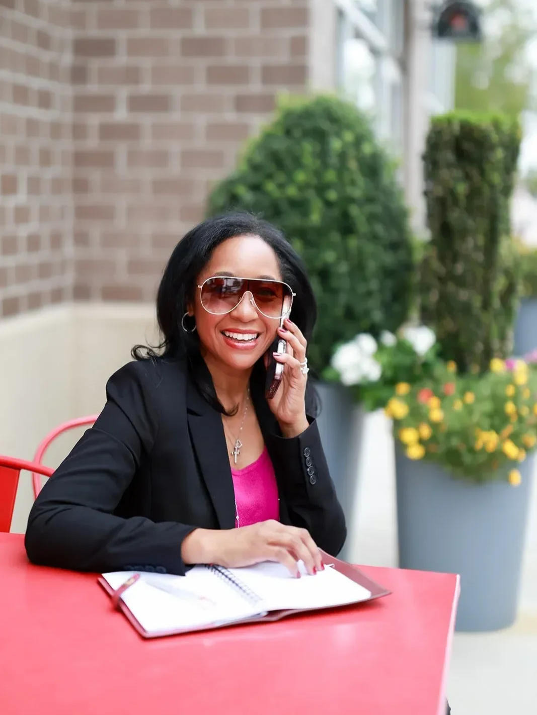 Dr. Betyshia Belardo sitting at a table outdoors, talking on a cellphone, smiling, wearing sunglasses, a black blazer, and a pink top, with an open notebook in front of her and plants in the background.