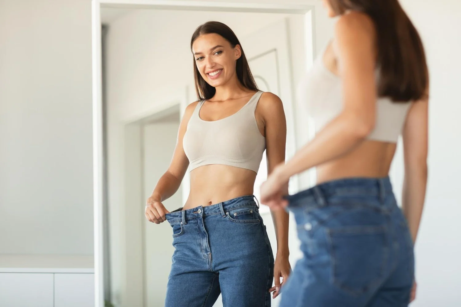 A woman looking in the mirror and smiling while trying on a pair of blue jeans.