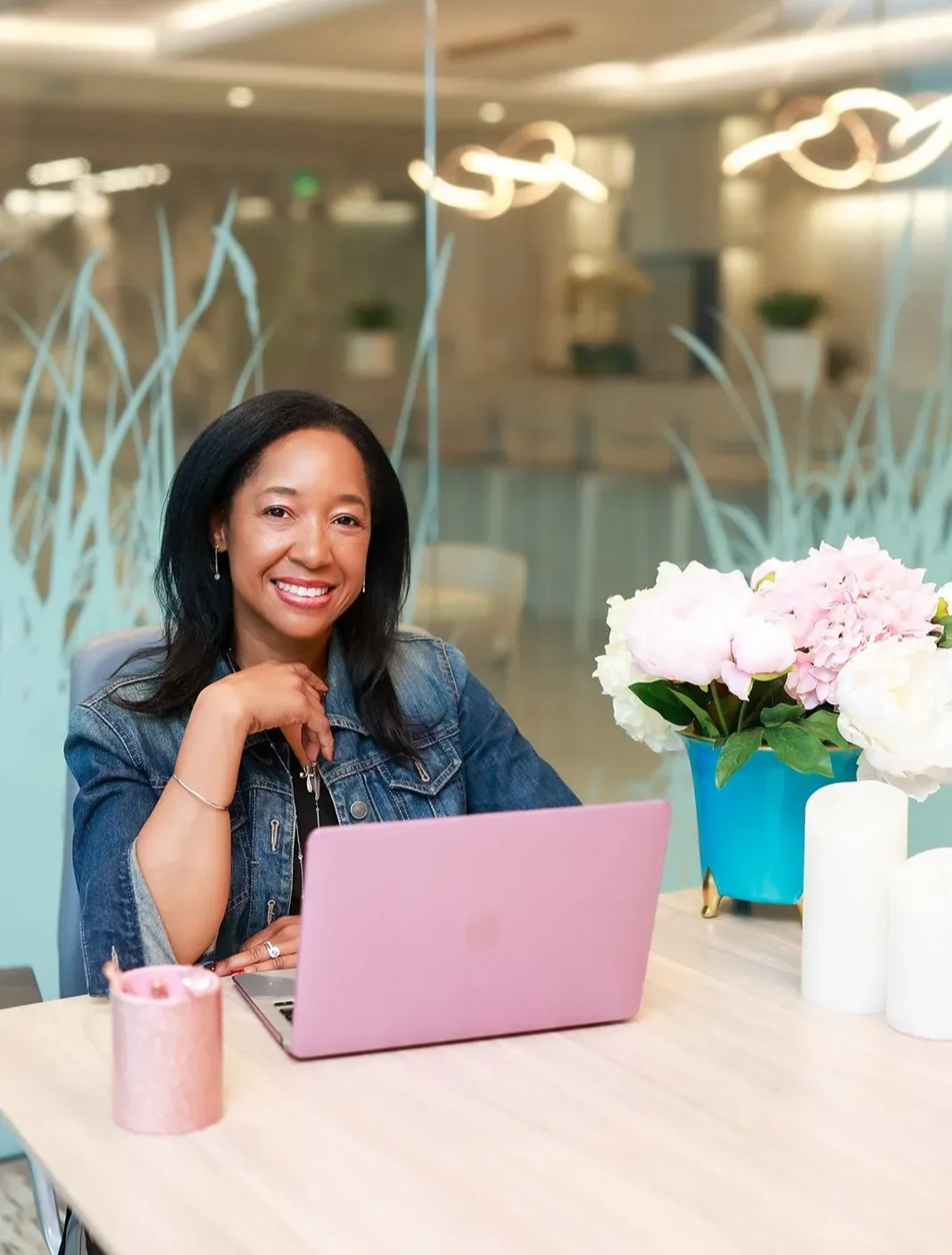 Dr. Betyshia Belardo sitting at a desk in an office, smiling, with a laptop and pink flowers on the desk, behind a glass wall with decorative patterns.