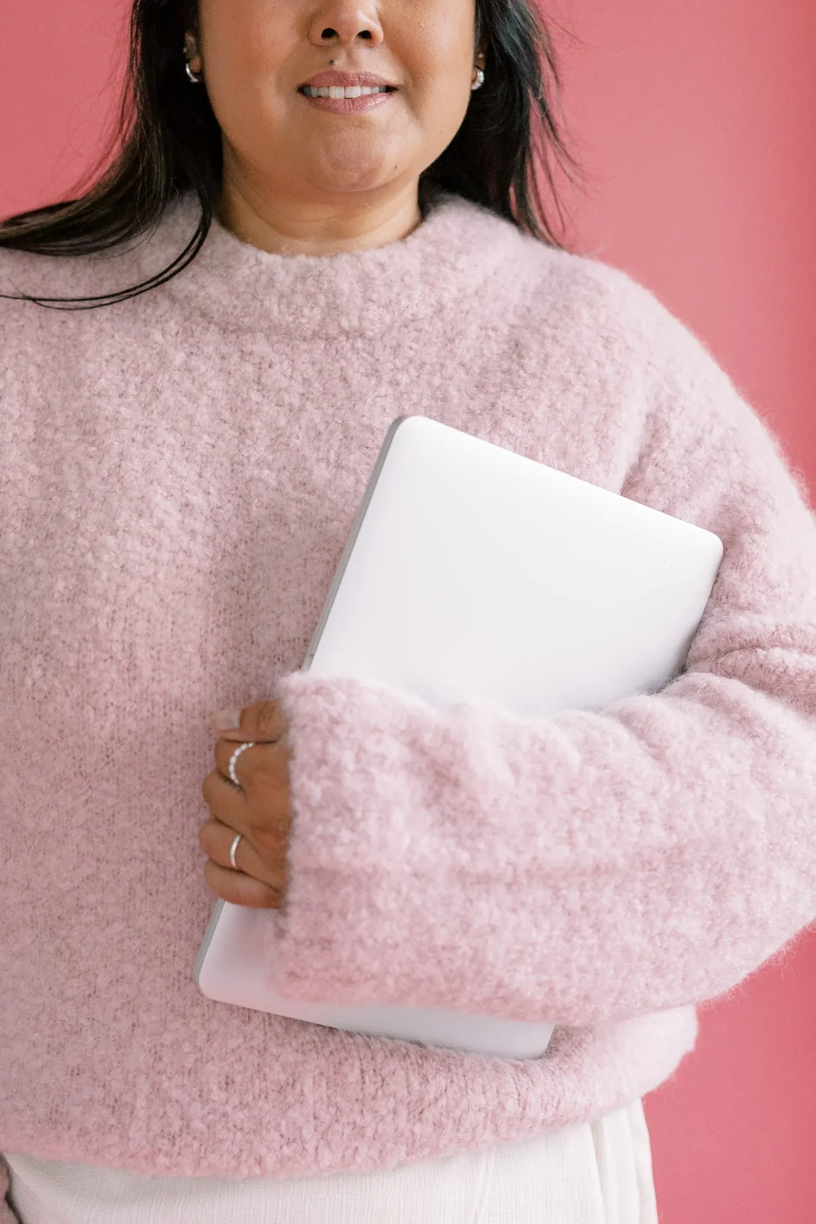 A woman wearing a pink, textured sweater and holding a closed white laptop, standing against a pink background.