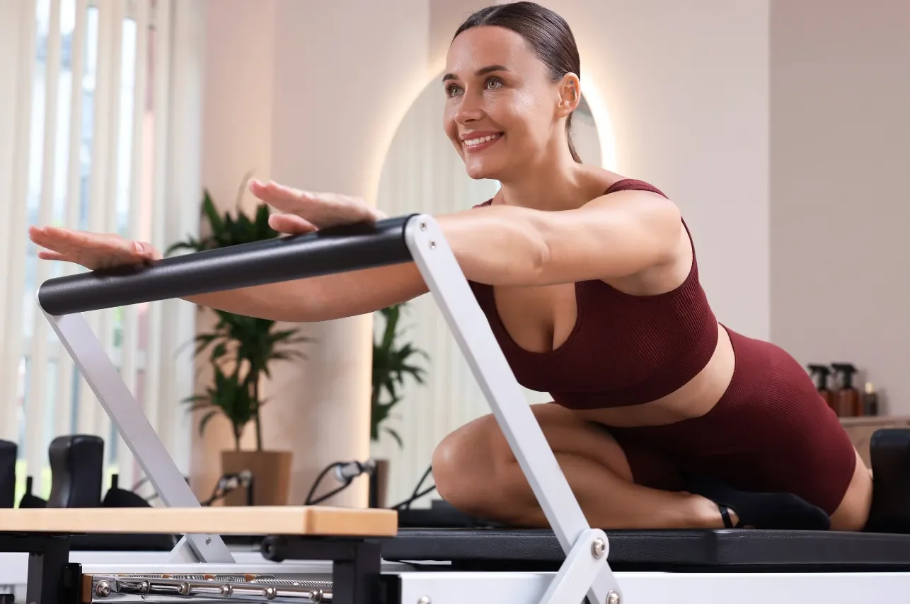 A woman practicing Pilates on a reformer machine in a fitness studio, smiling and wearing a matching maroon workout set.