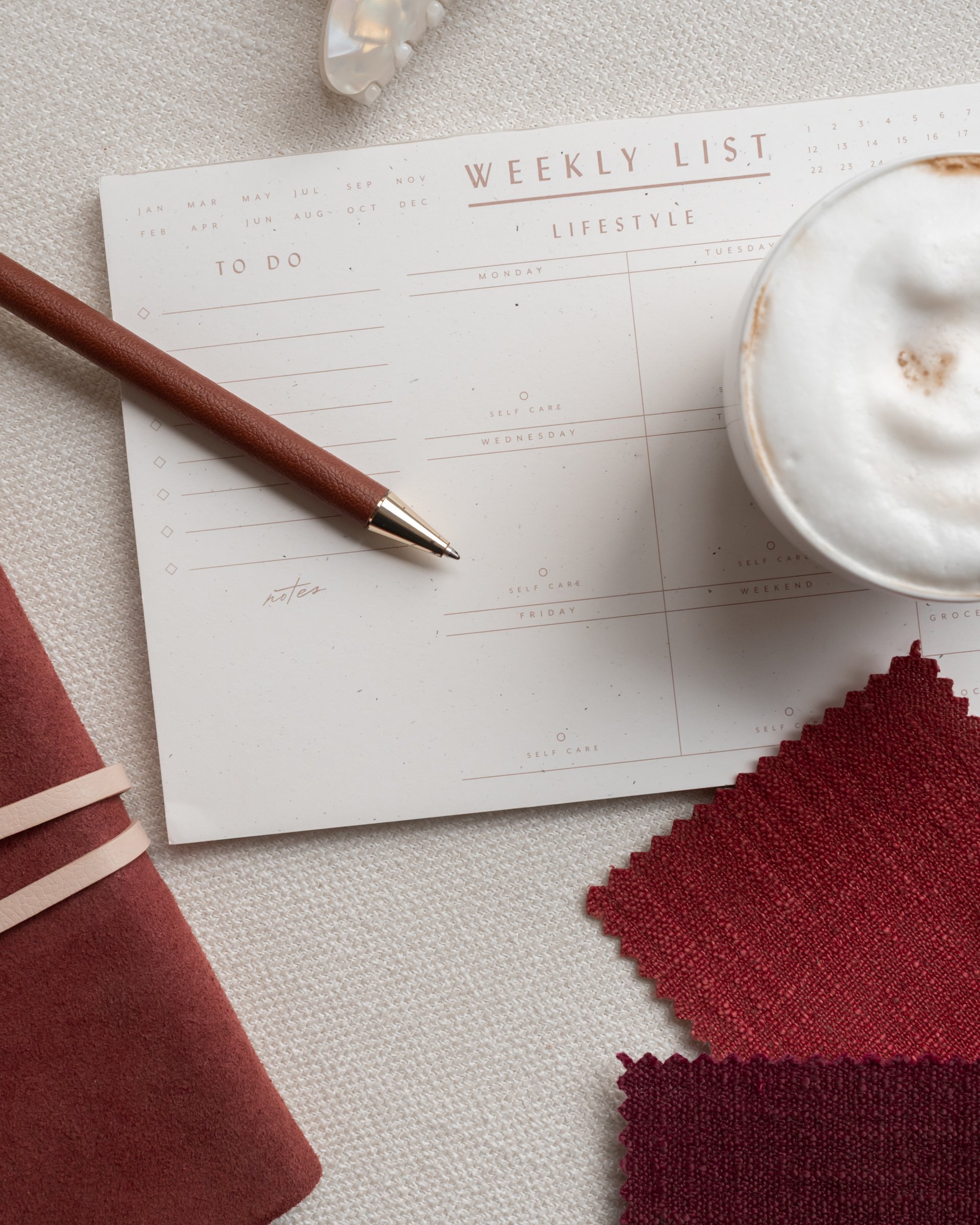 A weekly planner page with the days of the week, a maroon pen resting on the page, a cup of frothy coffee, and various red fabric swatches on a textured white surface.