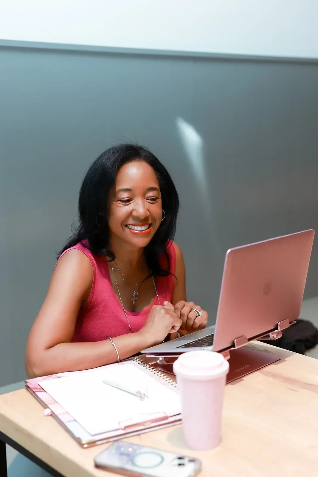 Dr. Betyshia Belardo wearing a pink sleeveless top, smiling and working on a pink laptop at a desk with notebooks, a pen, a pink insulated cup, and a smartphone.