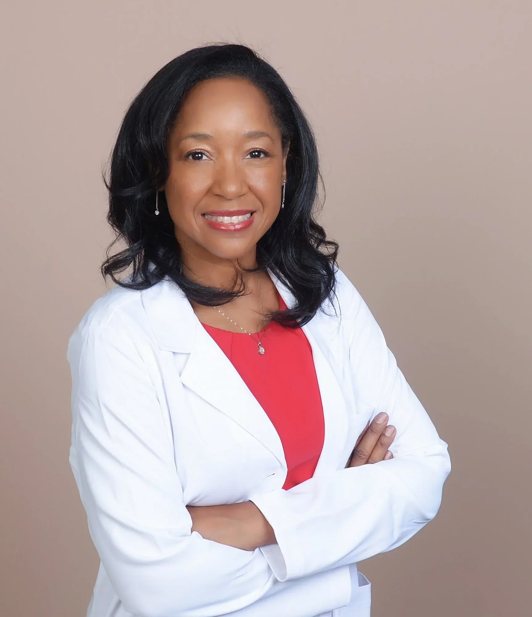 Dr. Betyshia Belardo with dark, curly hair wearing a white coat over a red top, smiling with arms crossed against a plain beige background.