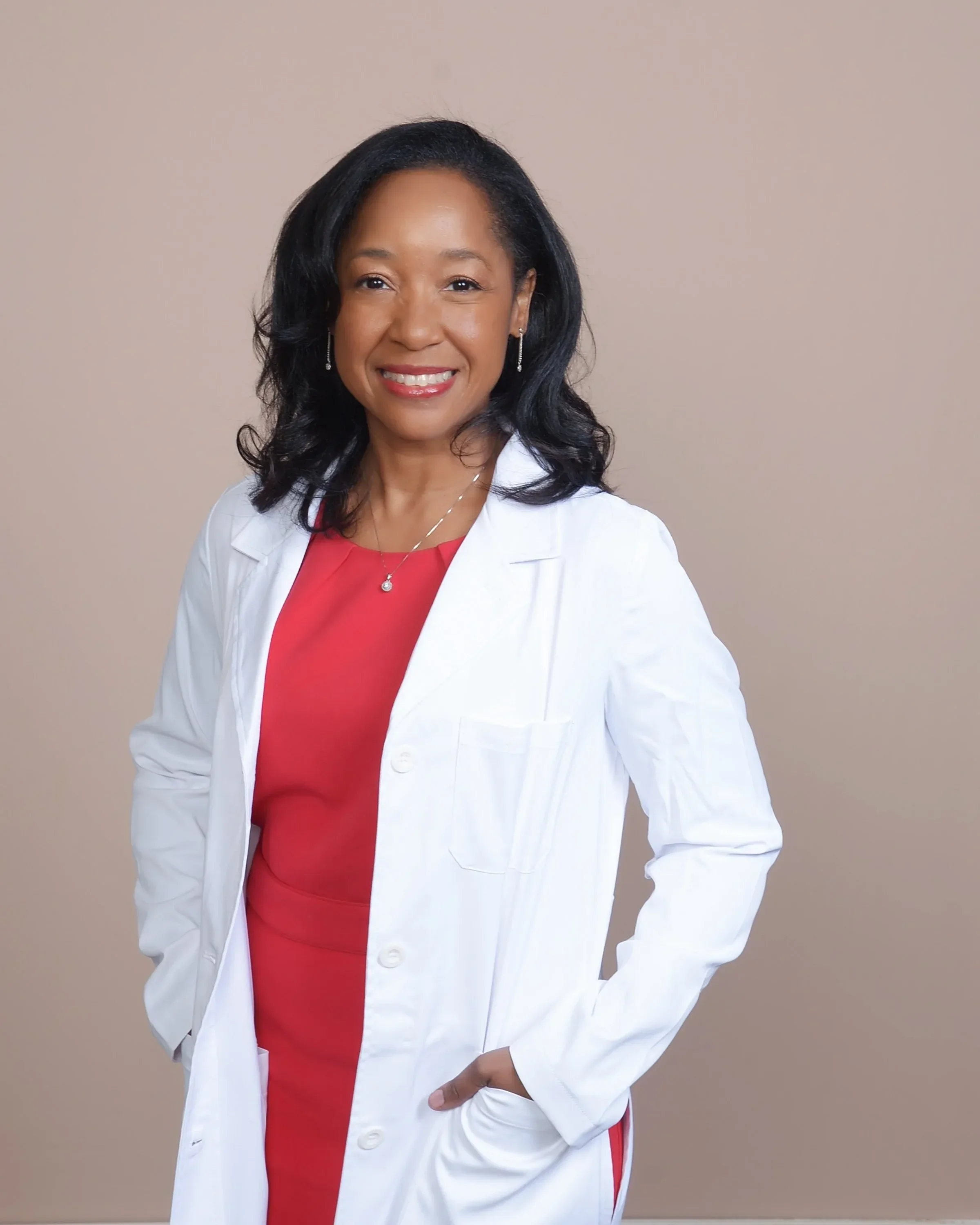 Dr. Betyshia Belardo wearing a white coat over a red top, with shoulder-length black hair and earrings, standing against a plain beige background.