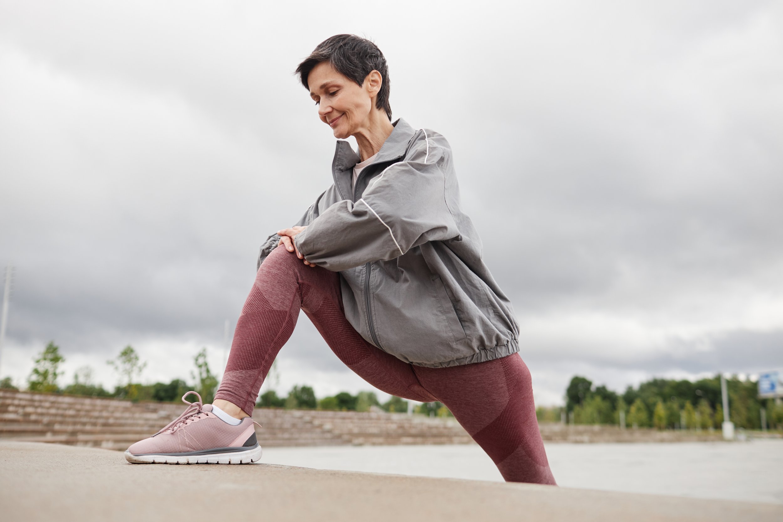 A woman stretching outdoors on a cloudy day, bending down with one leg forward and in a lunge position, wearing a gray jacket, pink leggings, and sneakers.