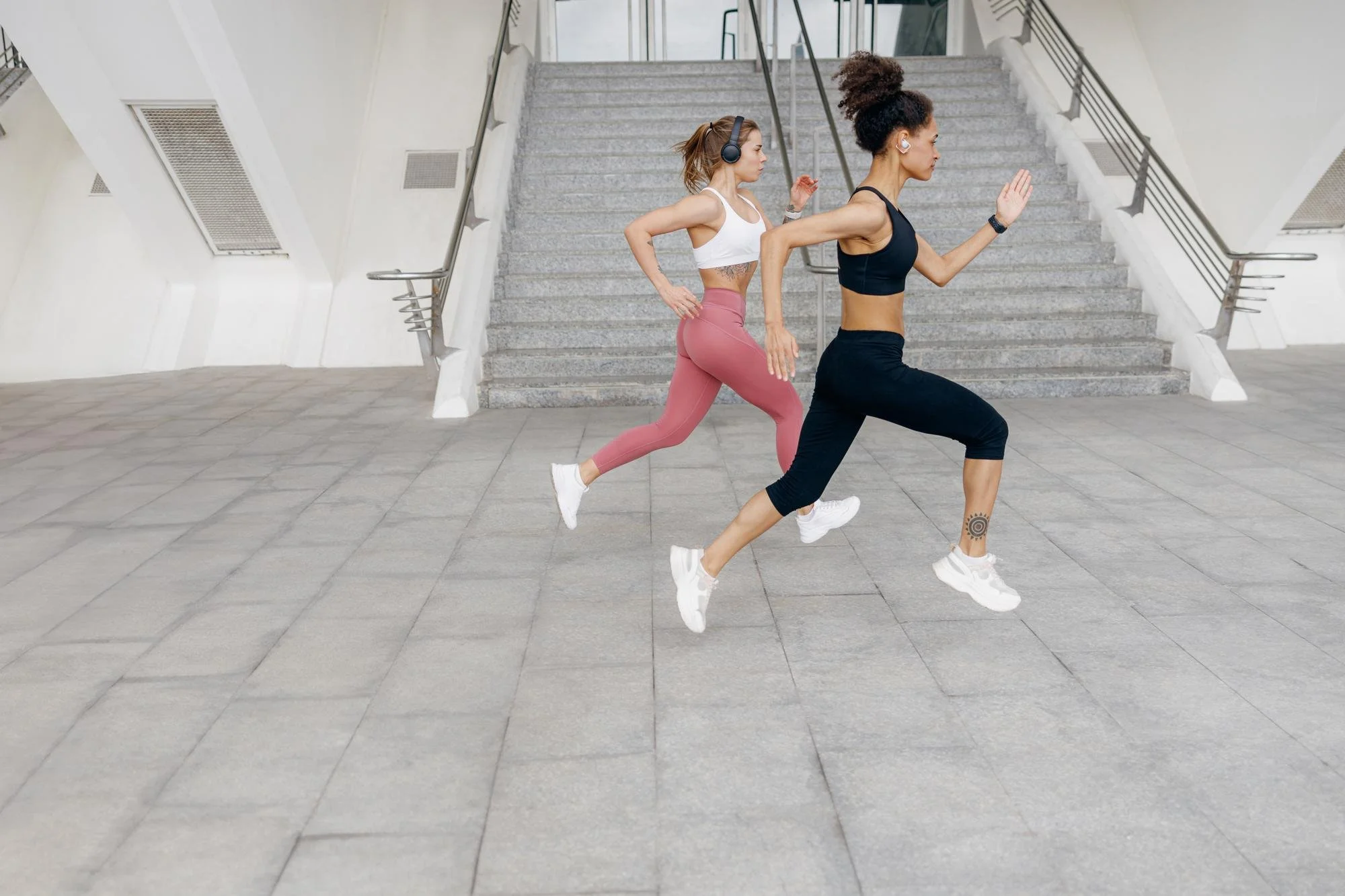 Two women running up a set of stairs in an indoor setting. One woman wears a black sports bra and black leggings, and the other wears a white sports bra and pink leggings. Both women are wearing wireless earbuds and athletic shoes.