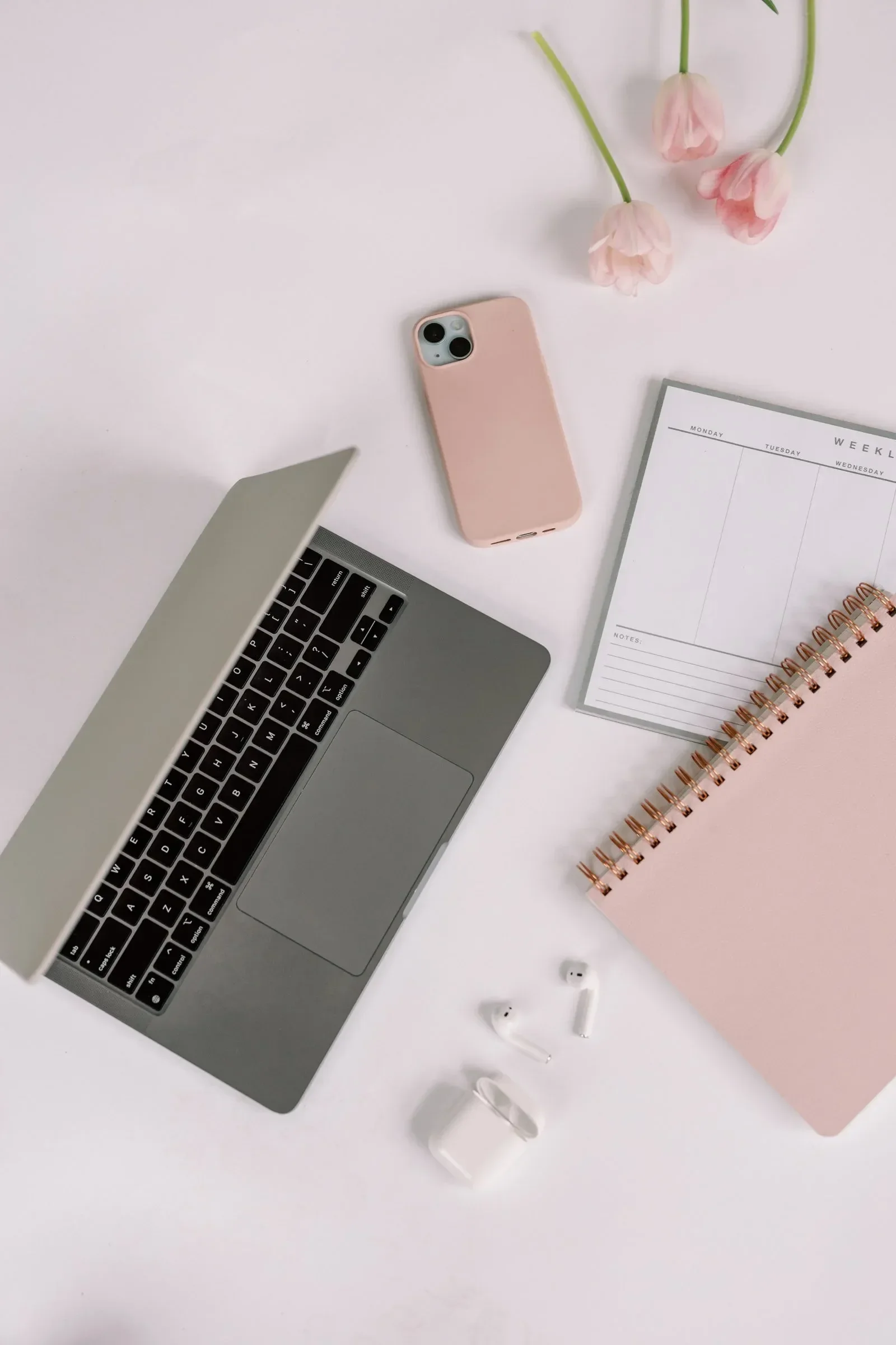 A flat lay of a workspace with a silver laptop, a pink smartphone, a white notebook with a spiral binding, pink tulips, a pair of white wireless earbuds, and a white wireless charging case on a white surface.